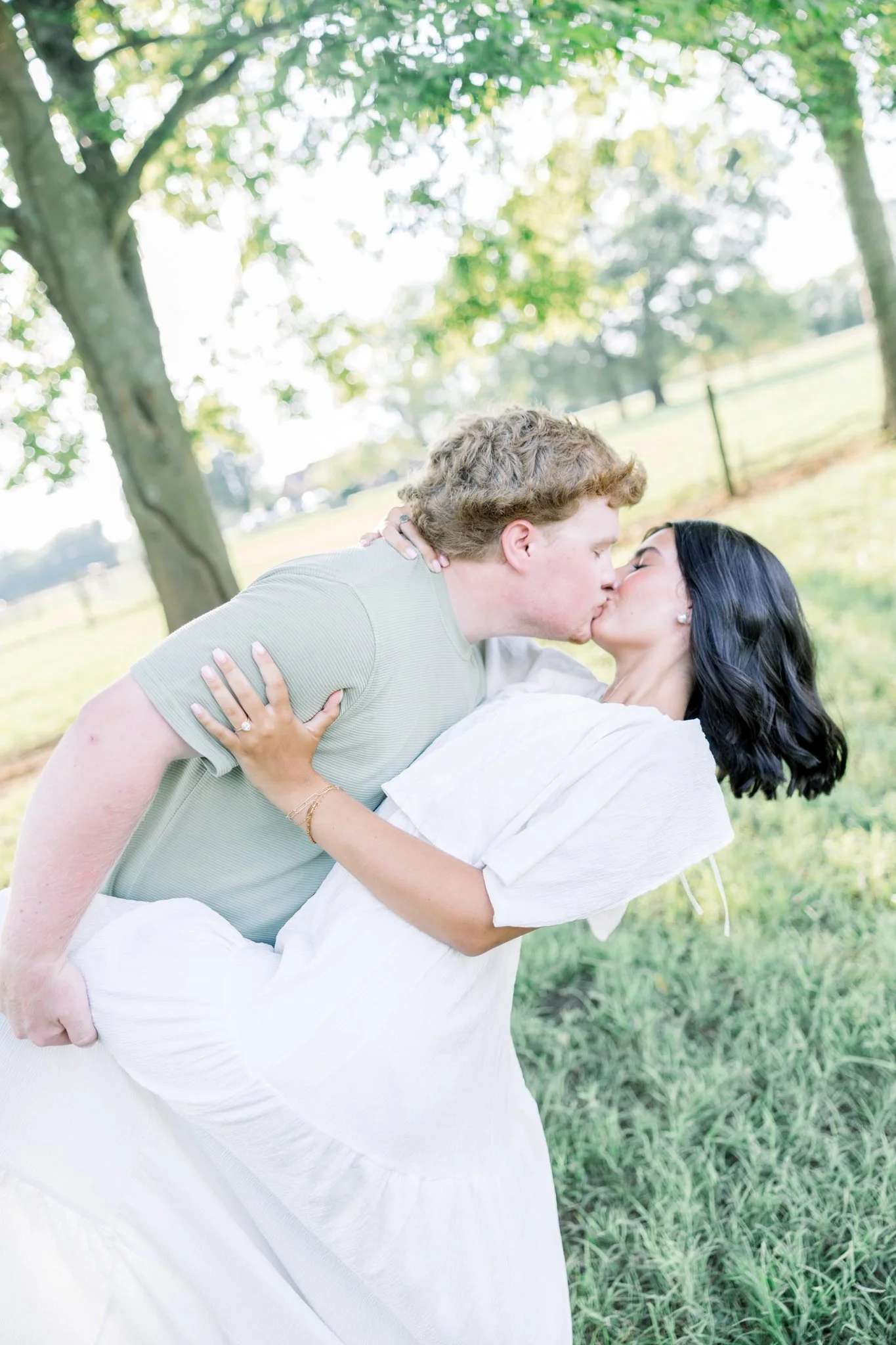 A couple is kissing outdoors in a park with green trees and grass in the background. The woman is wearing a white dress and has black hair, while the man is wearing a light green shirt and has curly blonde hair. The woman is holding the man, who is l