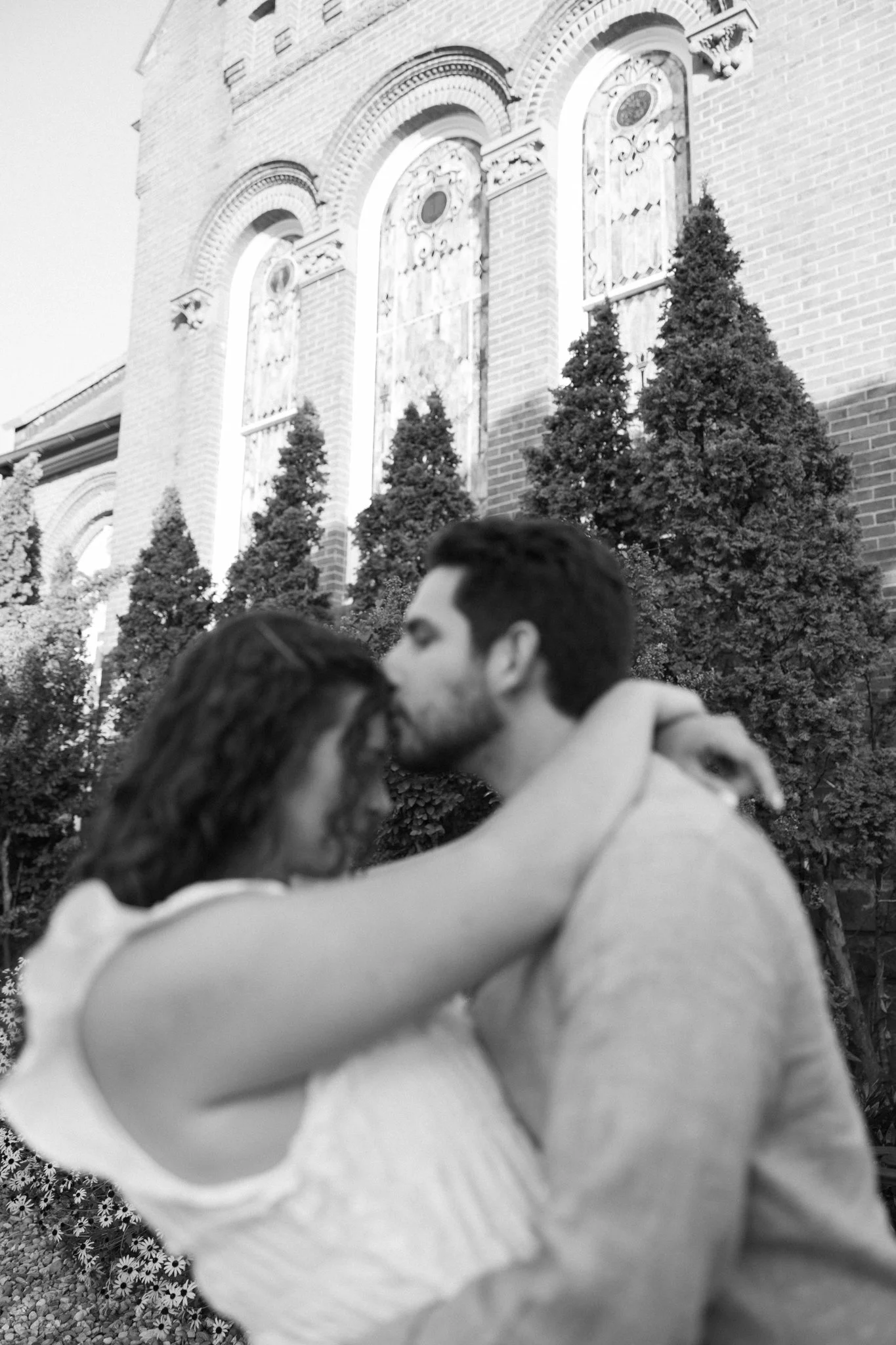 A couple hugging outdoors with a brick church and trees in the background, black and white photo.