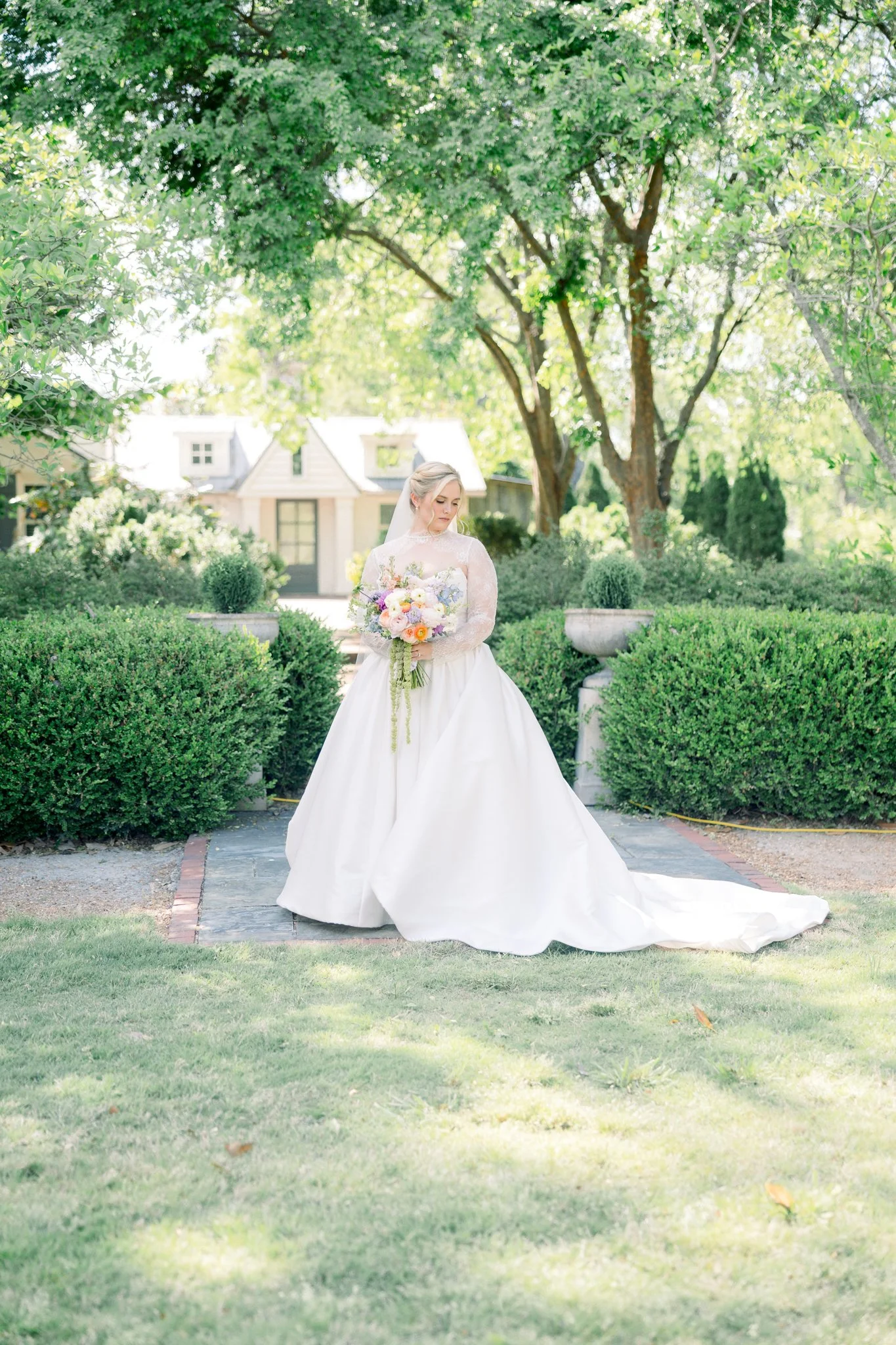 A bride in a white wedding gown holding a bouquet of flowers standing outdoors in a lush garden with green bushes and trees, and a small white house in the background.