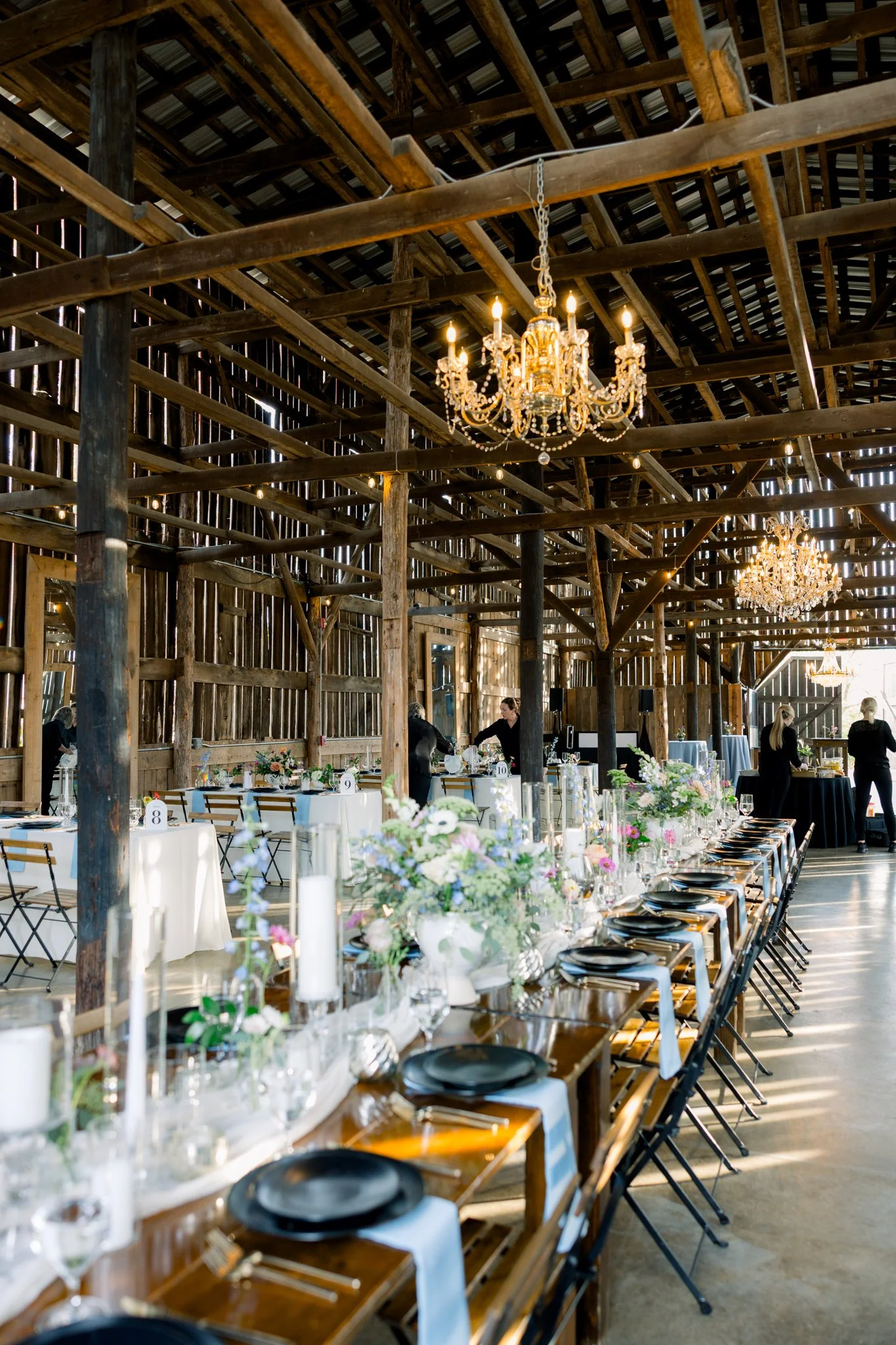 Interior of a rustic barn decorated for a wedding reception with long dining tables, white tablecloths, floral centerpieces, and black plates. Chandeliers hang from the wooden ceiling, and waitstaff are preparing the tables.