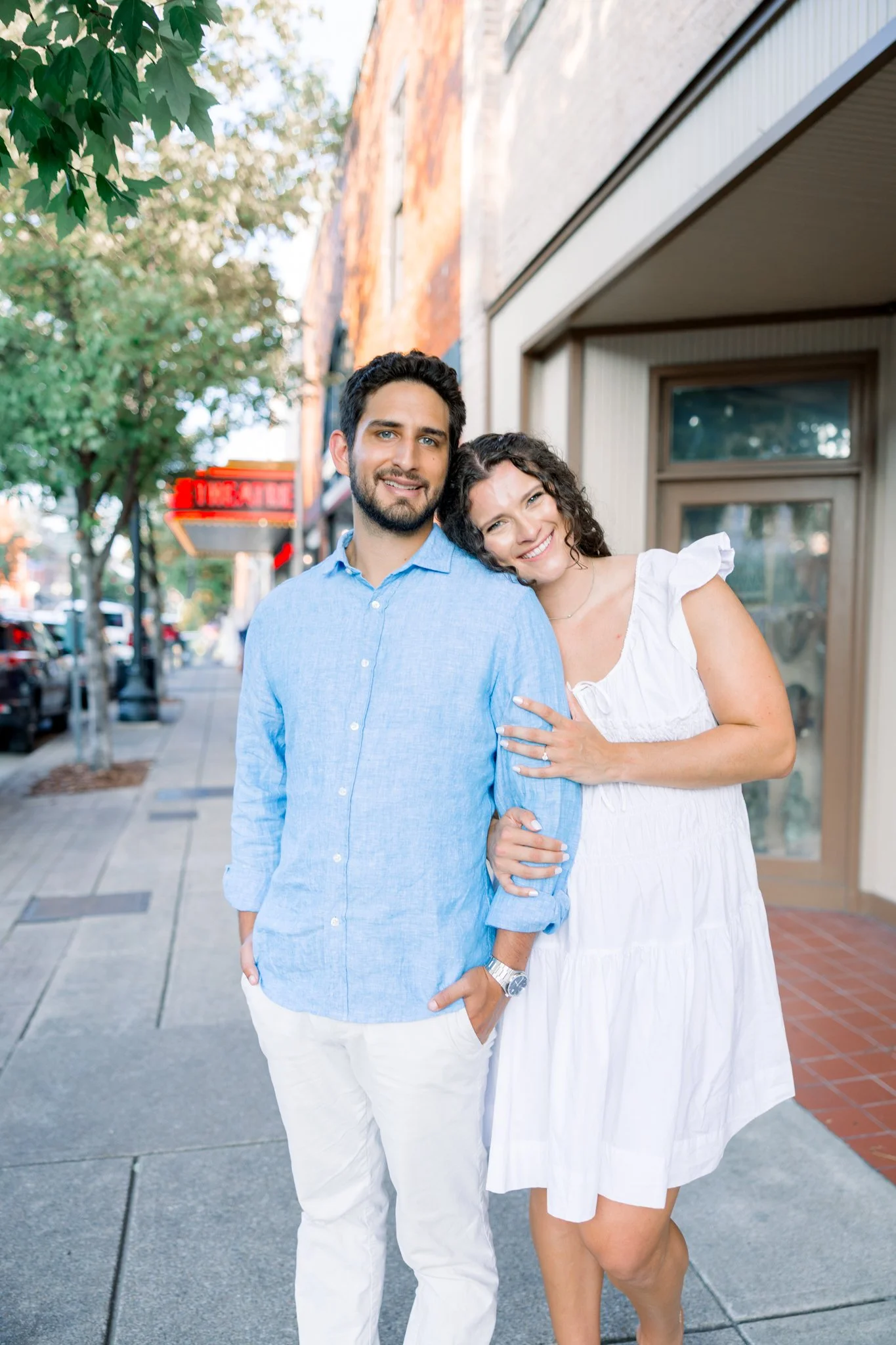 A young couple standing on a city sidewalk, smiling, with a man wearing a light blue shirt and white pants, and a woman in a white dress, leaning her head on his shoulder.