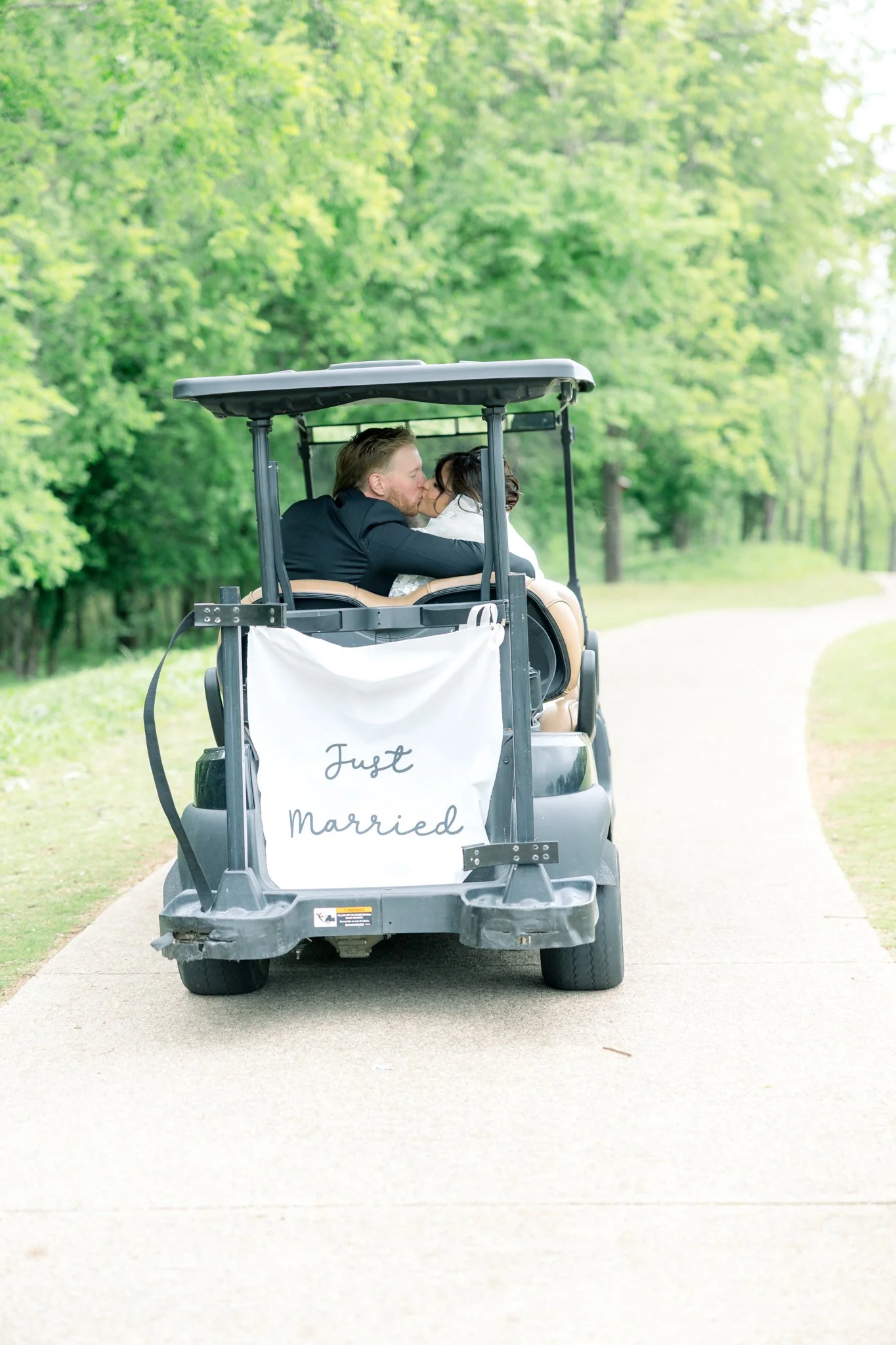 A newlywed couple kissing in a golf cart with a 'Just Married' sign, driving along a wooded park path.