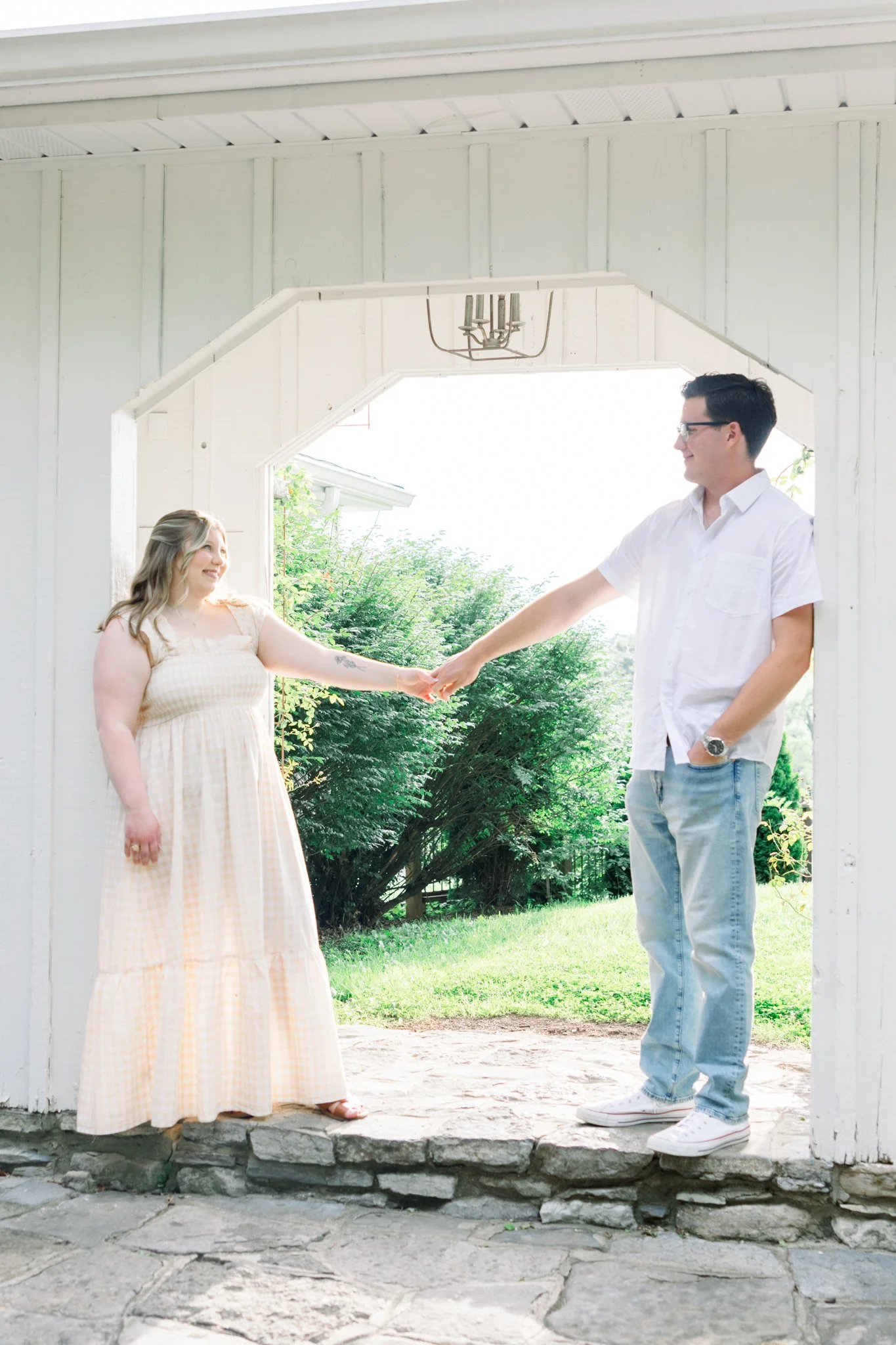 A young woman in a yellow checkered dress holding hands with a young man in a white shirt and jeans, standing under a white wooden porch with green bushes and trees in the background.
