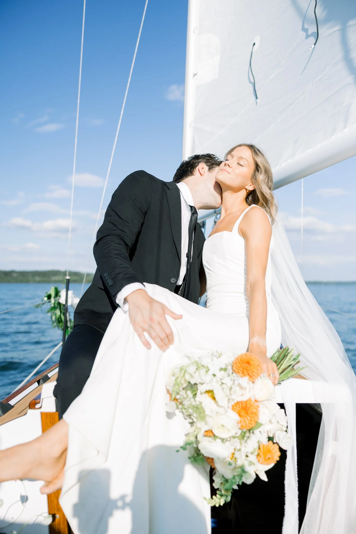A couple on a boat dressed in wedding attire, with the groom kissing the bride on the cheek. The bride holds a bouquet of white, orange, and green flowers, with the water and blue sky in the background.