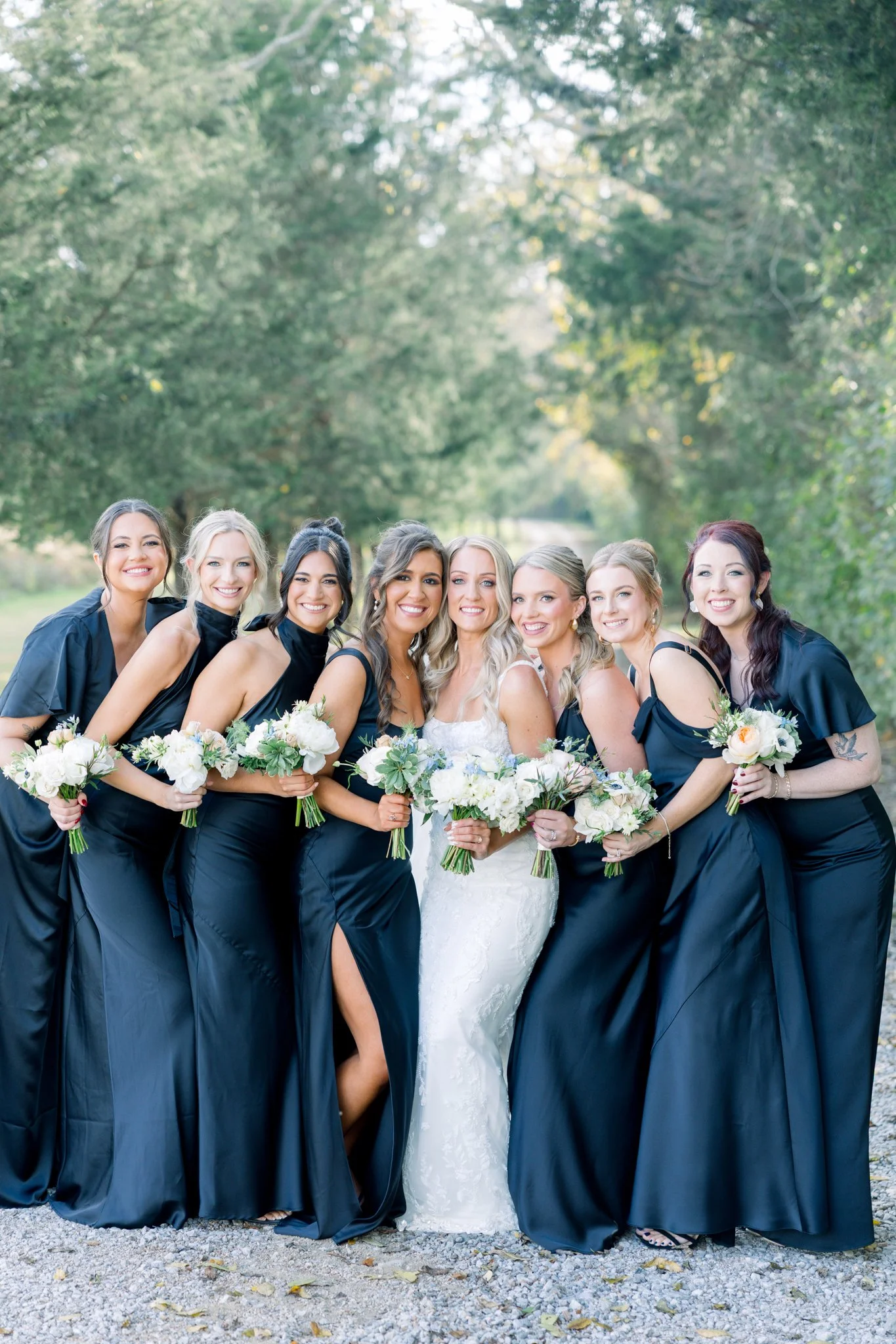 A bride in a white wedding dress standing amidst her bridesmaids dressed in black gowns outdoors, all holding bouquets of white flowers and smiling.