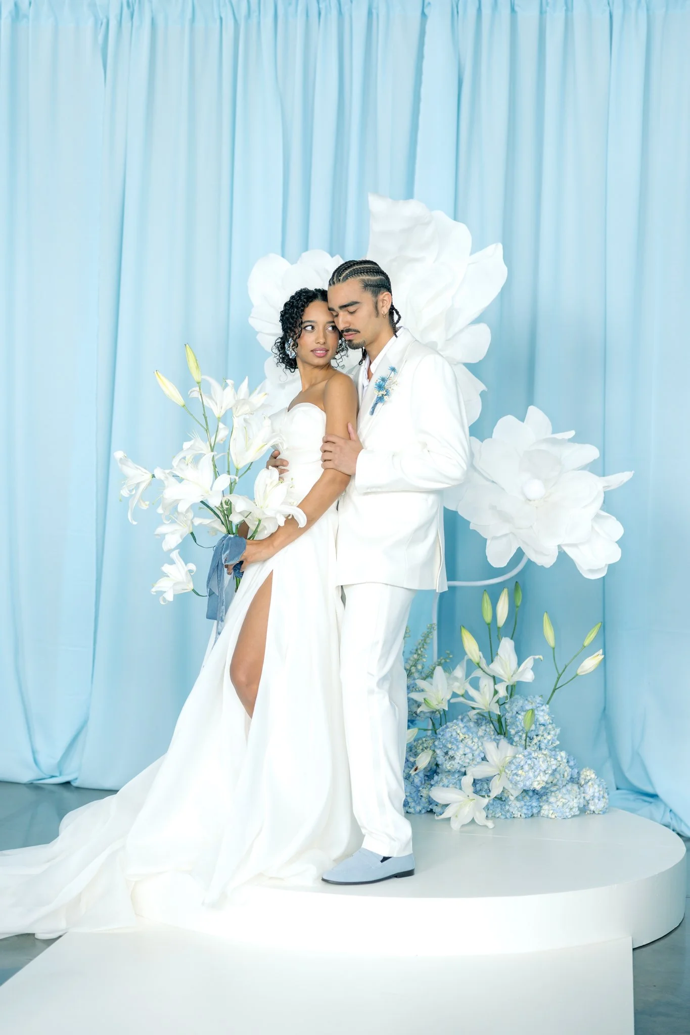 A couple dressed in white wedding attire posing on a platform decorated with white and blue flowers, with a backdrop of light blue curtains.