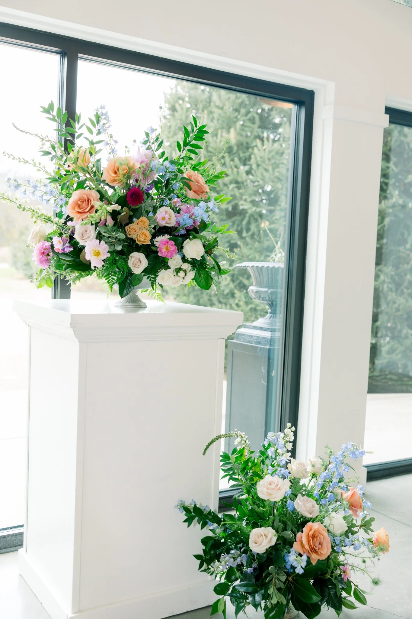A floral arrangement with pink, white, and purple flowers on a white pedestal near large glass windows with trees outside.