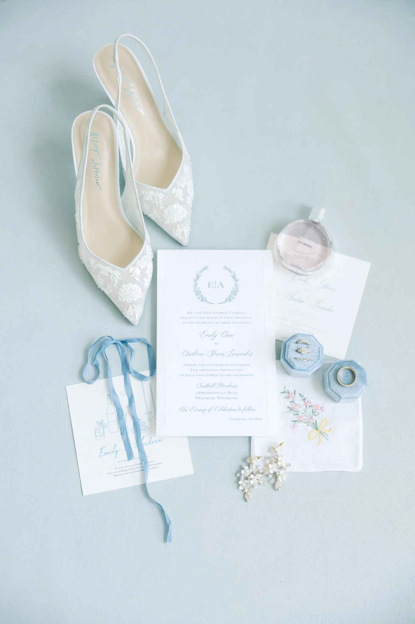 White bridal shoes with floral embroidery, a perfume bottle, wedding invitation cards, a ring, jewelry, and decorative elements arranged on a light blue surface.