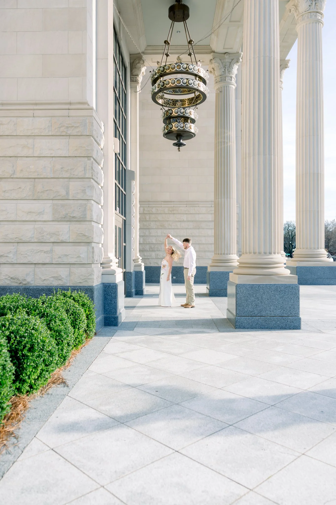 A couple dressed for a wedding, standing on steps in front of a neoclassical building with large columns, a chandelier hanging from the ceiling, and sky visible in the background.