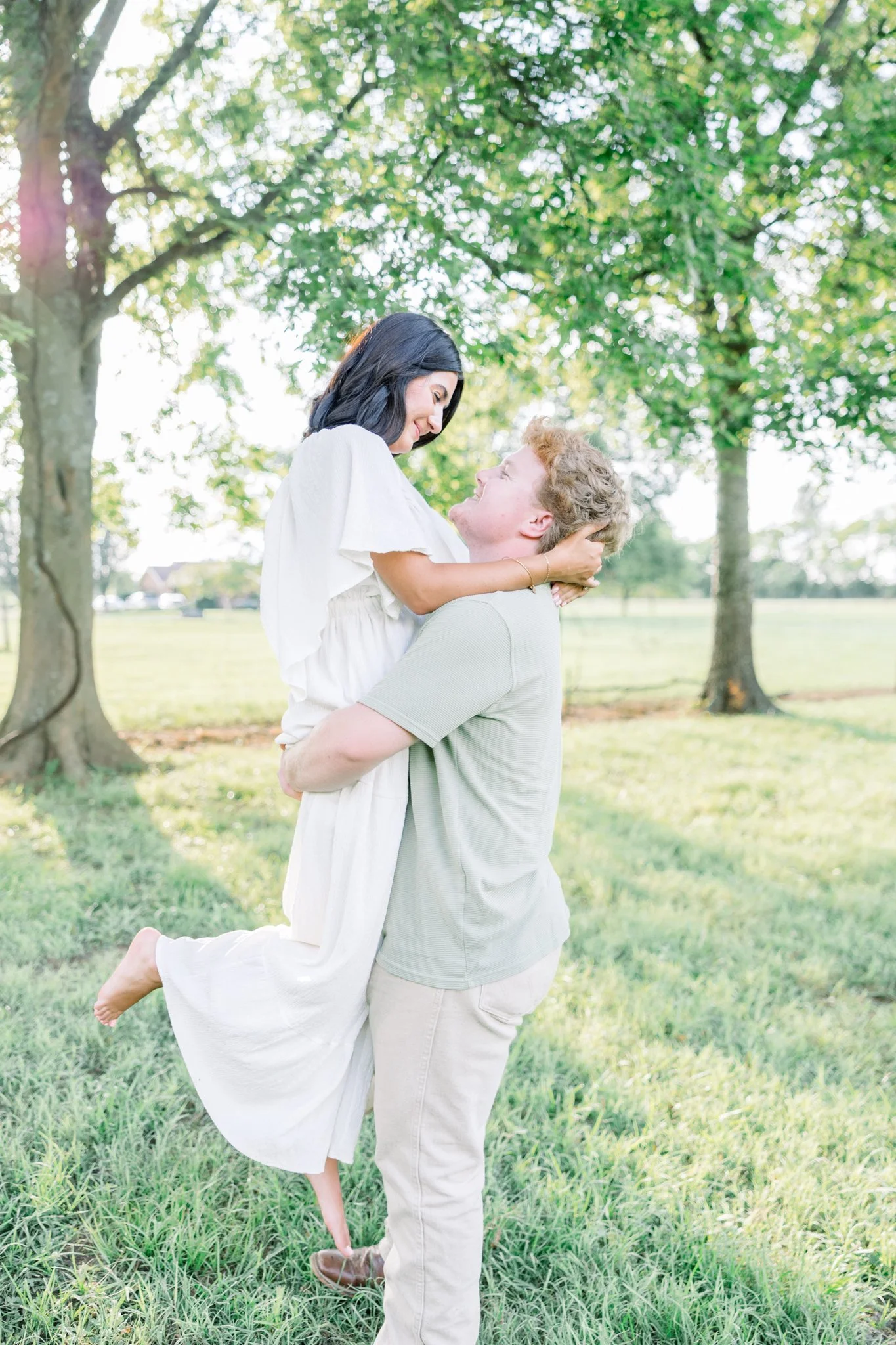 A couple outdoors, the man holds the woman in his arms, both smiling and looking at each other, surrounded by trees and green grass during daytime.