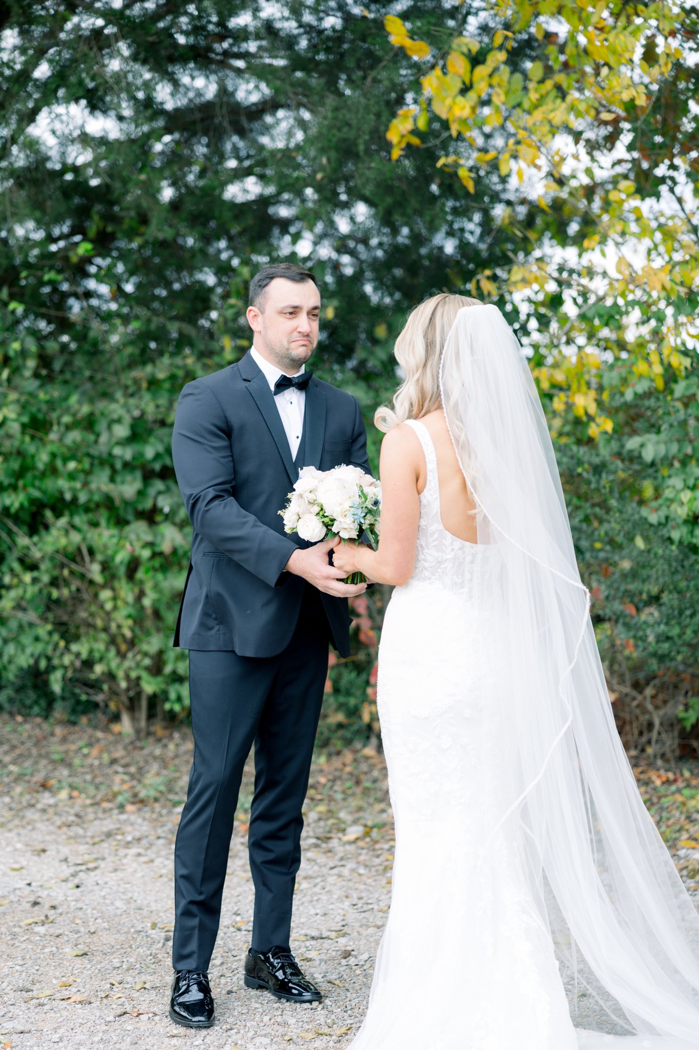 A groom in a black tuxedo with a bowtie is holding hands with a bride wearing a white wedding dress with lace details and a long veil. The groom has a sad expression on his face, while the bride's face is not visible. They are outdoors with green tre