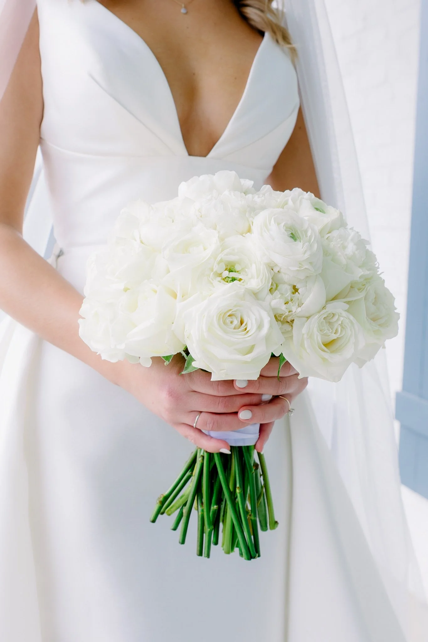 A bride in a white dress holding a bouquet of white roses and lisianthus flowers. The focus is on the bouquet and upper body, with a plain background.