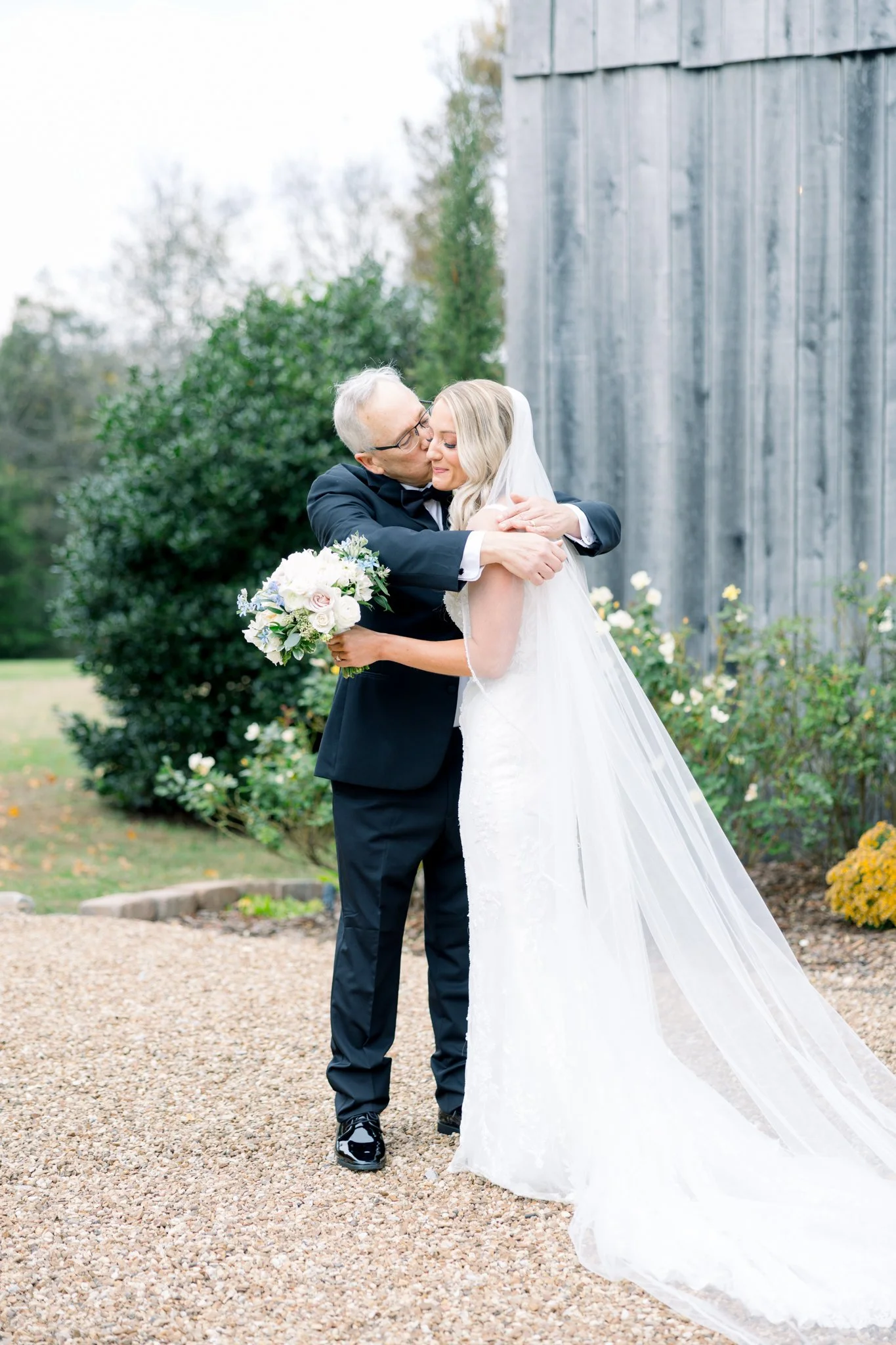 An elderly man in a tuxedo embracing a young bride in a white wedding gown outdoors, holding a bouquet of flowers, with a rustic wooden building and greenery in the background.