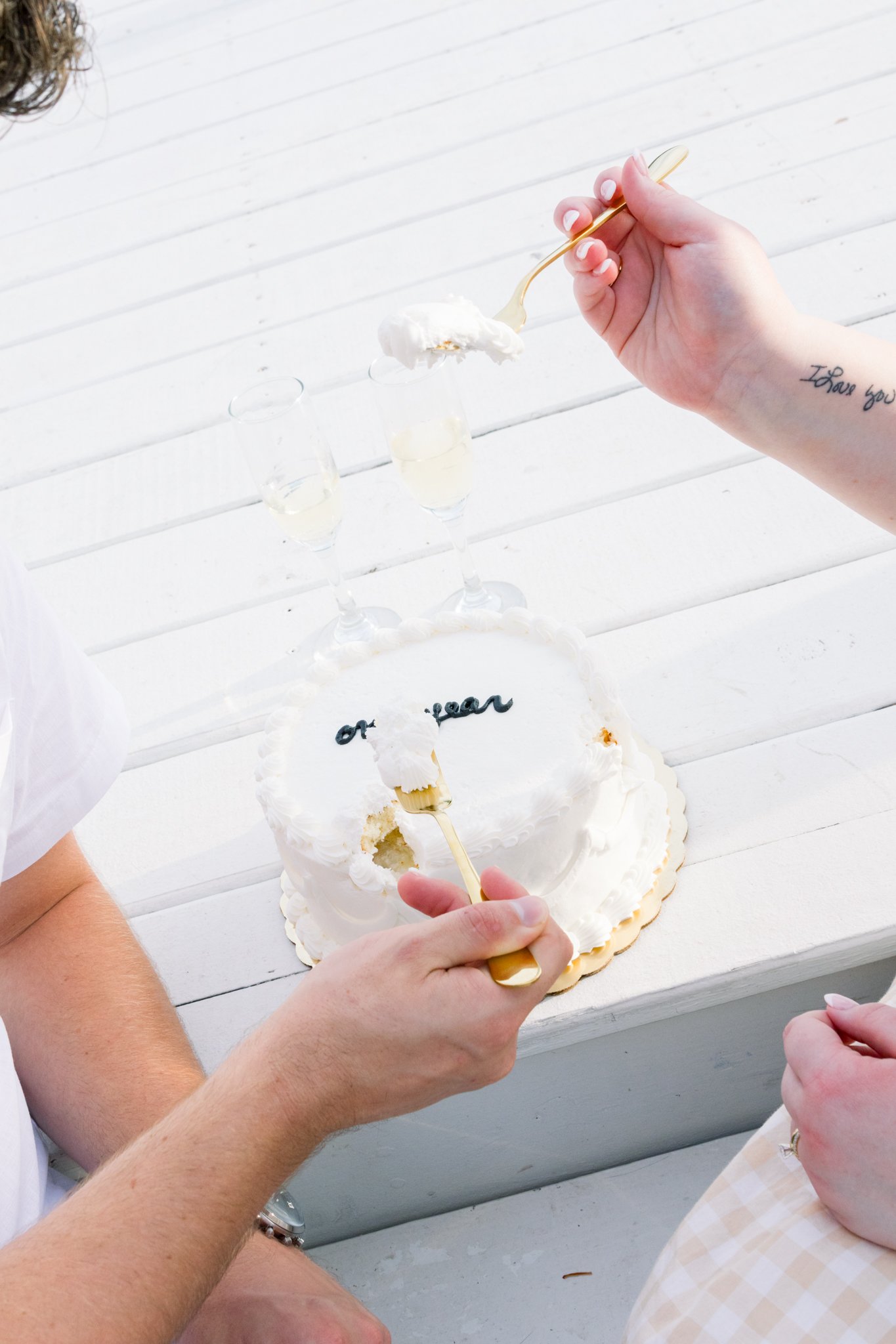 Person holding a spoonful of whipped cream over a white cake with the words 'of your' written on it, set on a white wooden table.