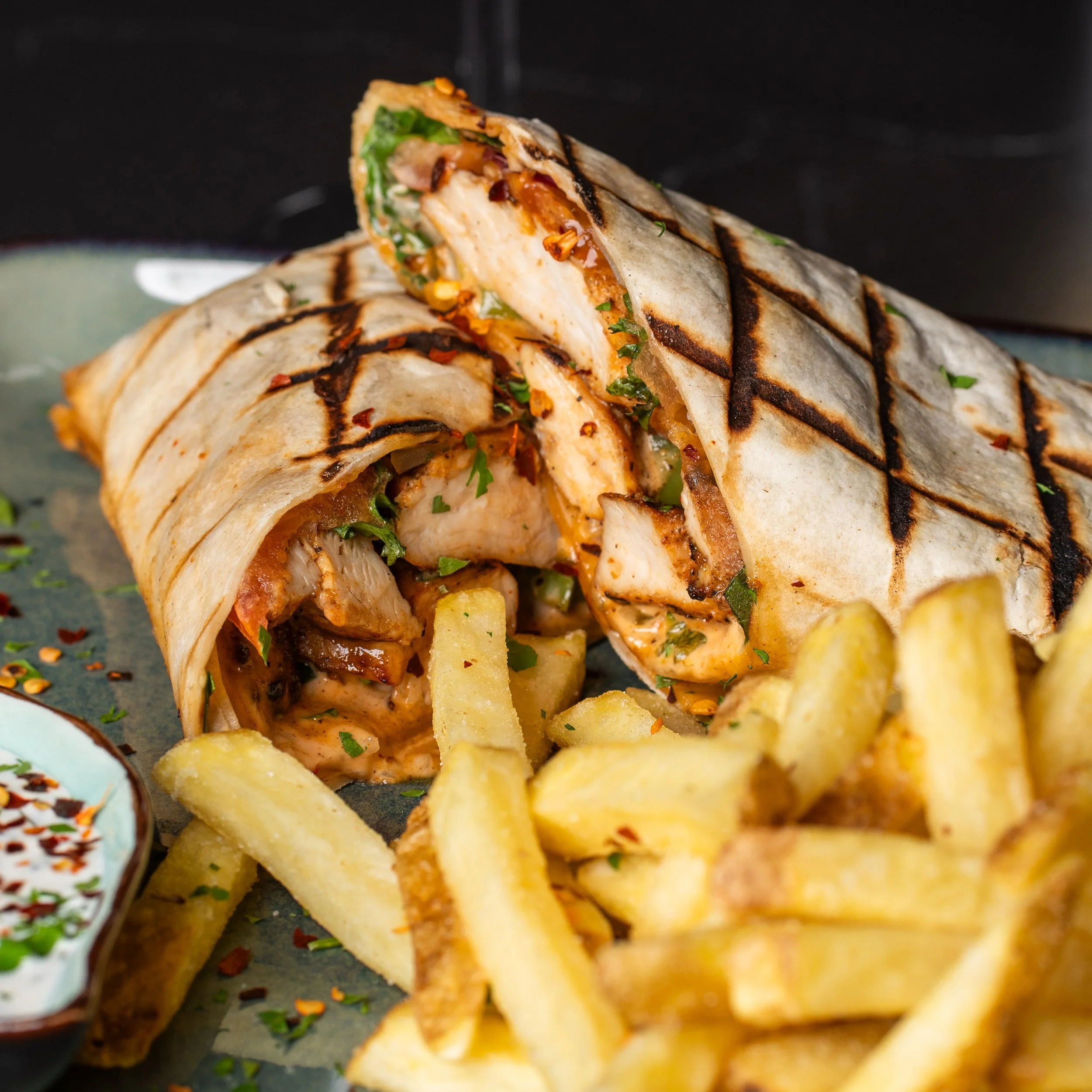 Close-up of a grilled wrap filled with chicken, vegetables, and sauce, served with potato chips and garnished with herbs.