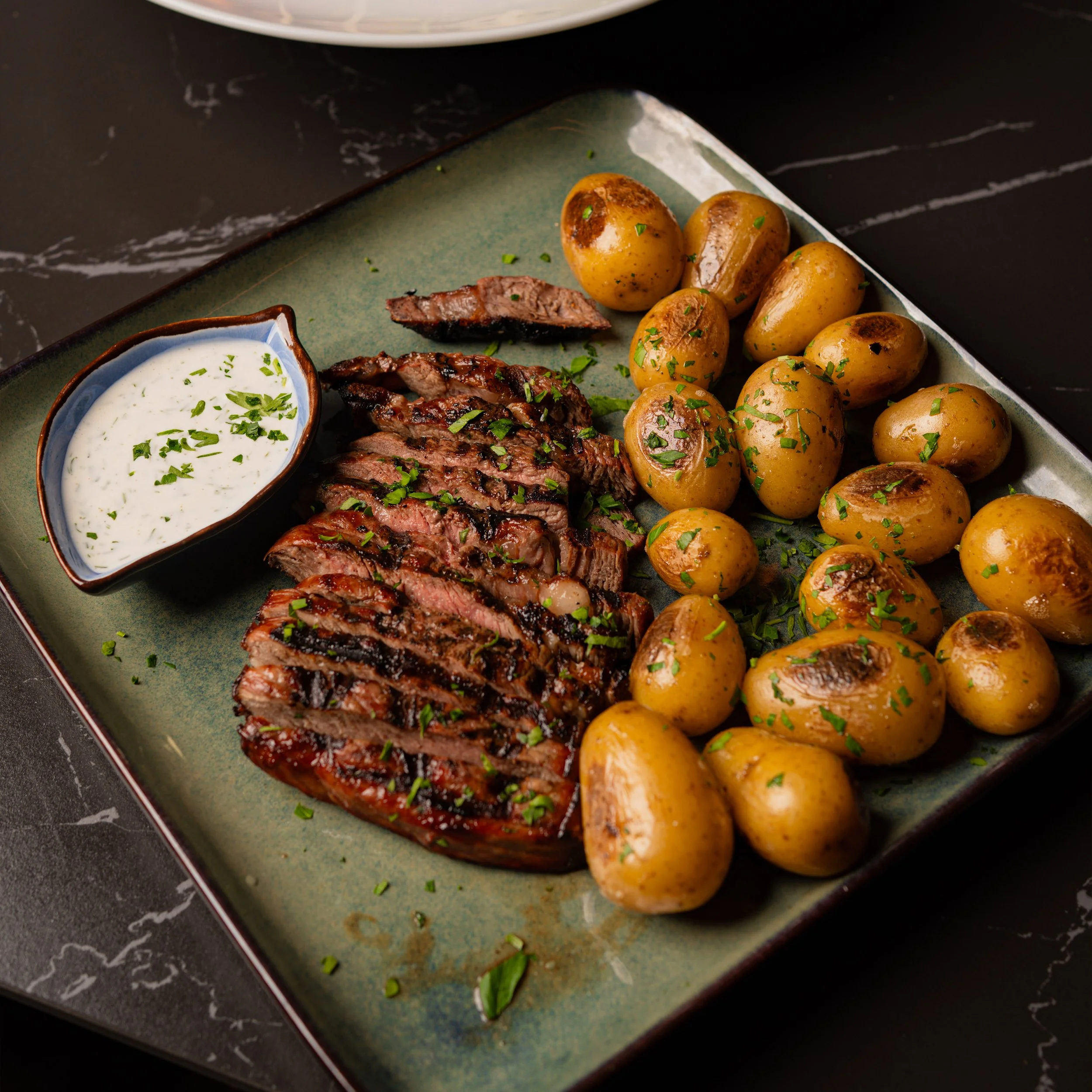 A plate of grilled steak, roasted potatoes garnished with herbs, and a small bowl of creamy sauce with herbs on a green plate.