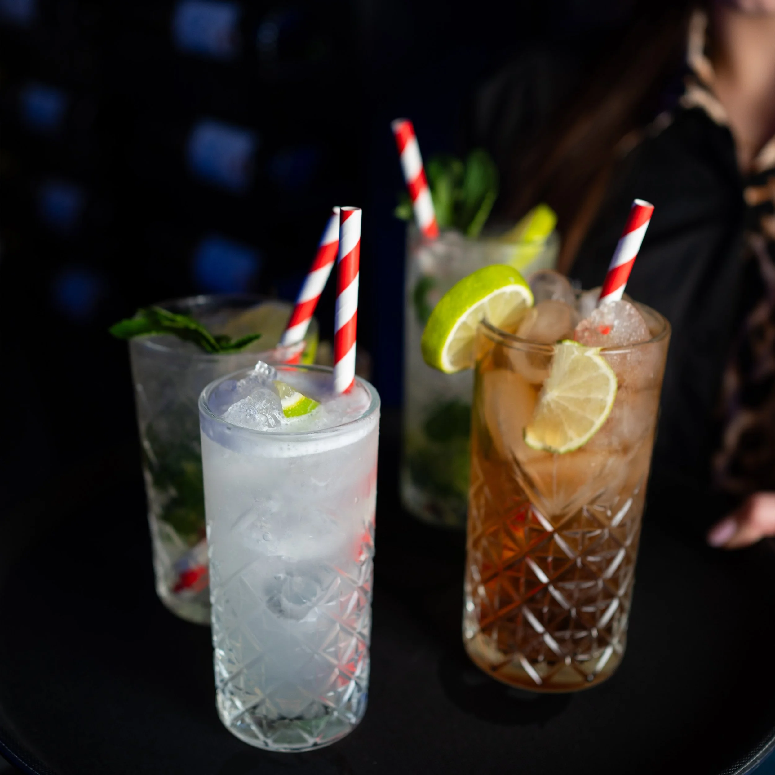 Close-up of cocktails with lemon slices and striped straws on a dark surface.