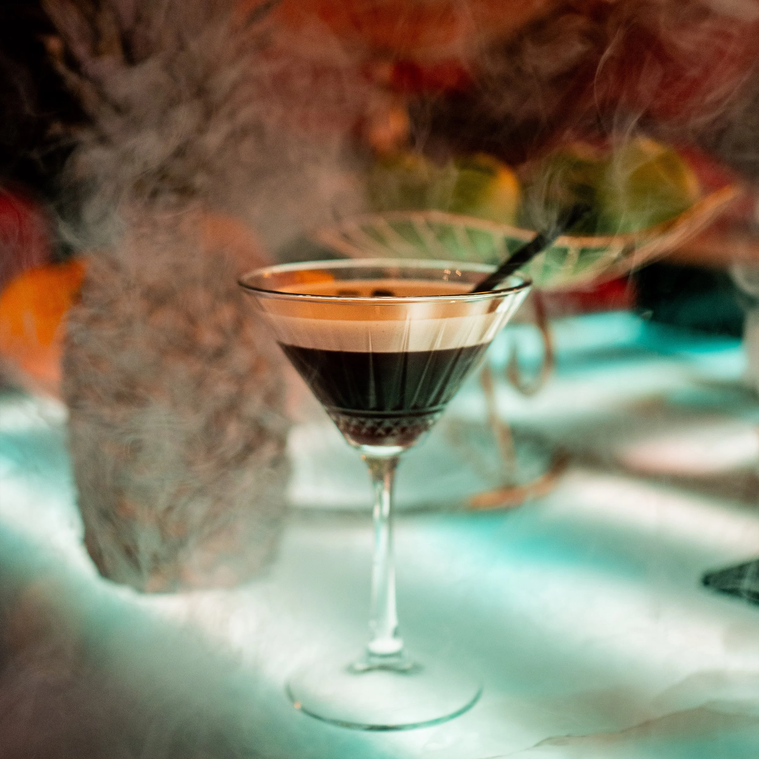 A close-up of a martini glass filled with a dark liquid, with steam or smoke rising from the drink, against a blurred background with warm tones.
