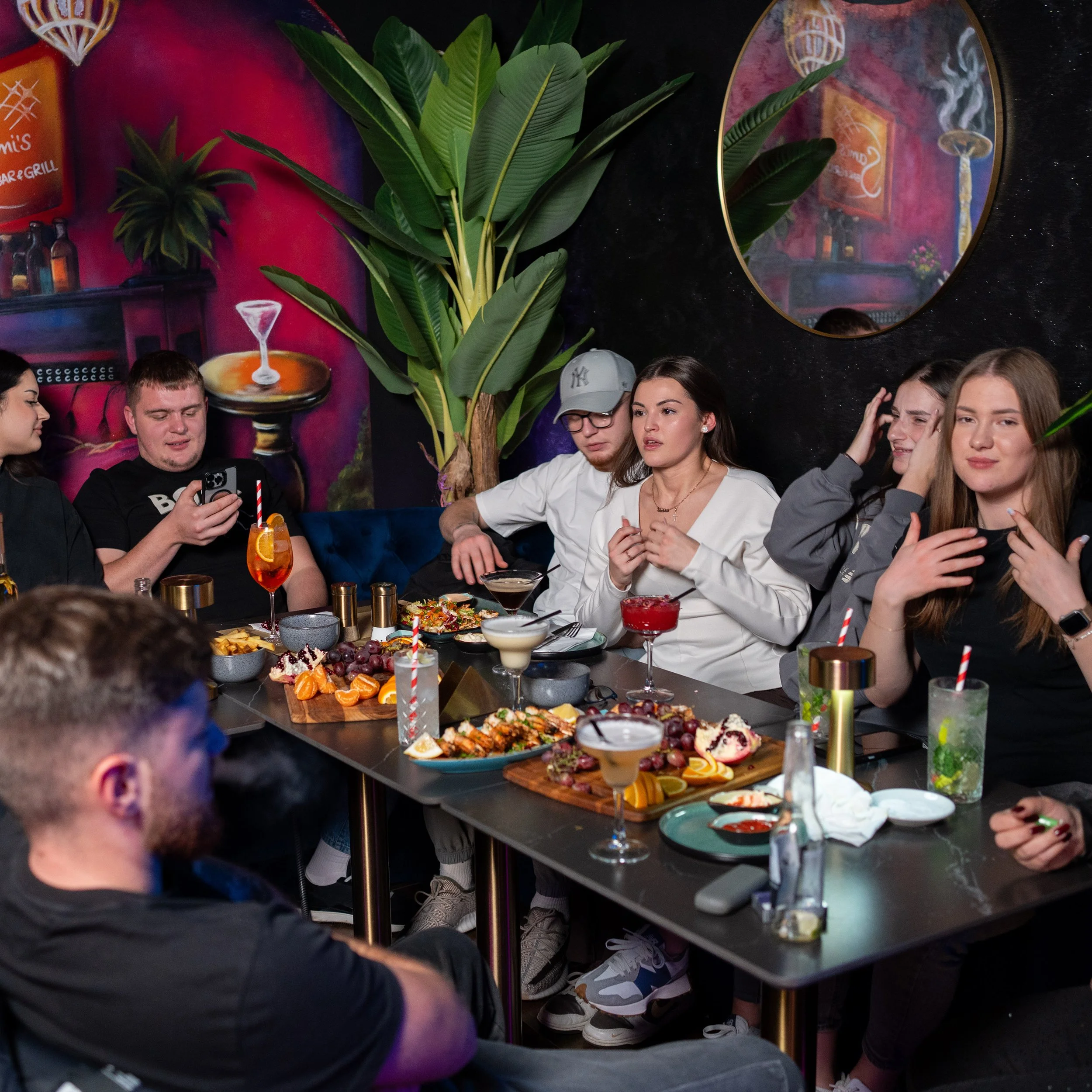 A group of young people gathered around a table with various dishes of food and drinks, including cocktails and fruit platters, in a restaurant or bar setting with colorful wall art and a large decorative mirror.