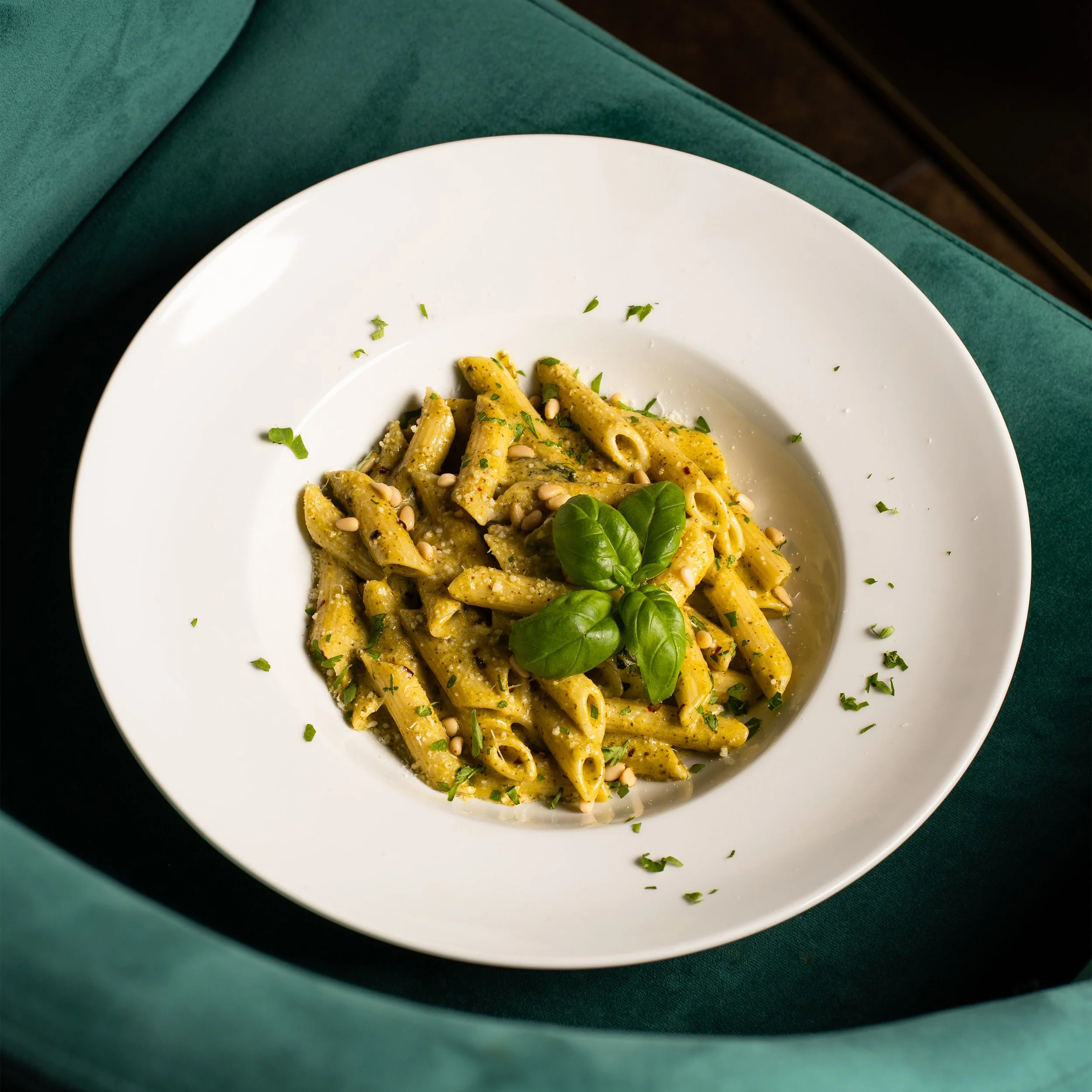 A white plate of pasta with green pesto sauce, garnished with chopped herbs, pine nuts, and fresh basil leaves, on a dark green tablecloth.