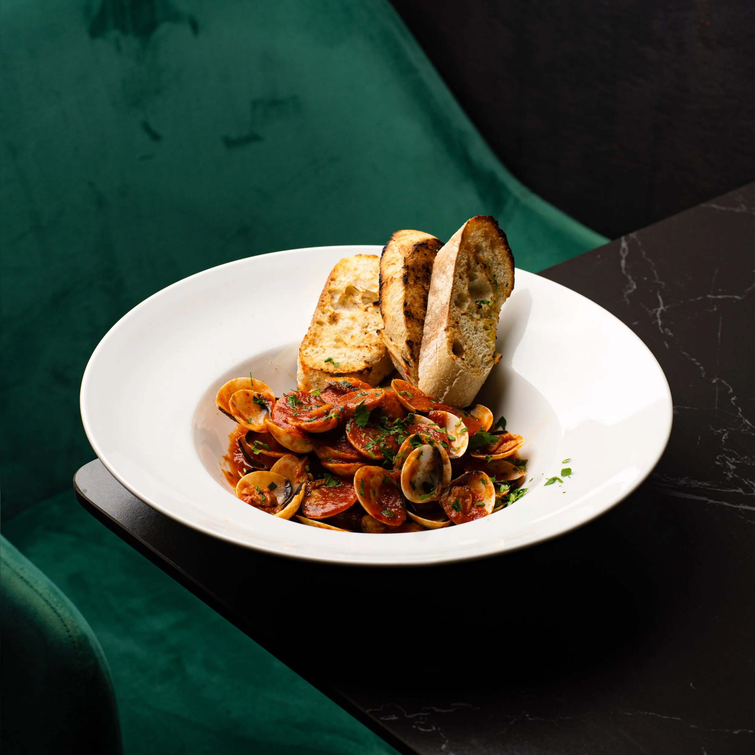 A white bowl containing a pasta dish with tomato sauce, topped with chopped herbs, and slices of toasted bread on the side, set on a dark marble surface with a green wall in the background.