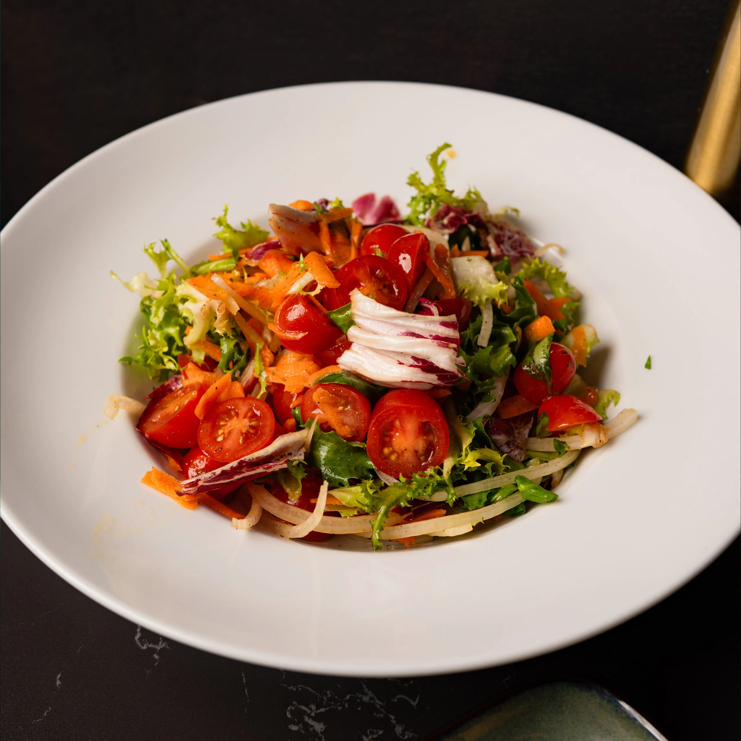 Close-up of a fresh vegetable salad with cherry tomatoes, shredded carrots, lettuce, and radicchio in a white bowl on a black surface.
