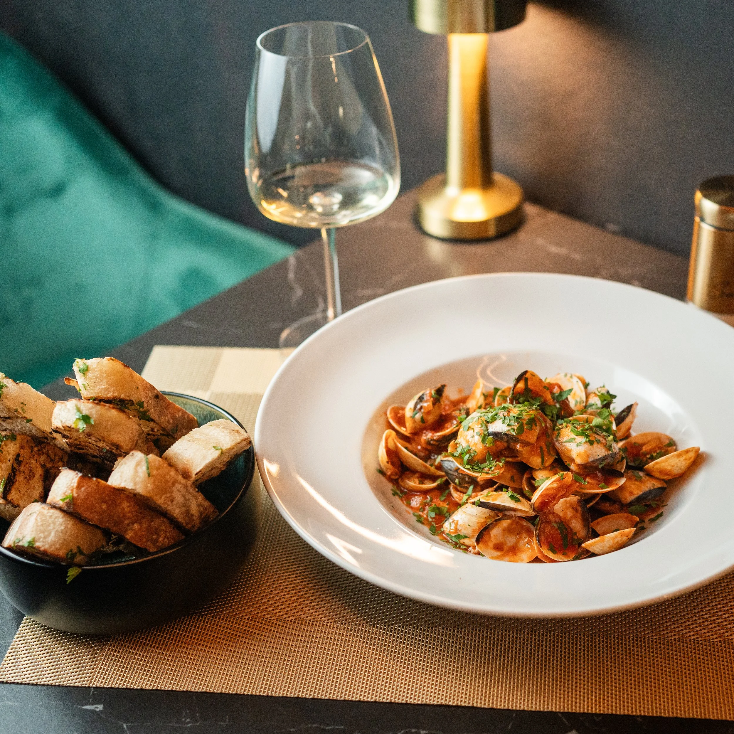A table setting with a glass of water, a gold lamp, a bowl of seafood pasta garnished with parsley, slices of toasted bread with herbs, and a napkin on a placemat.