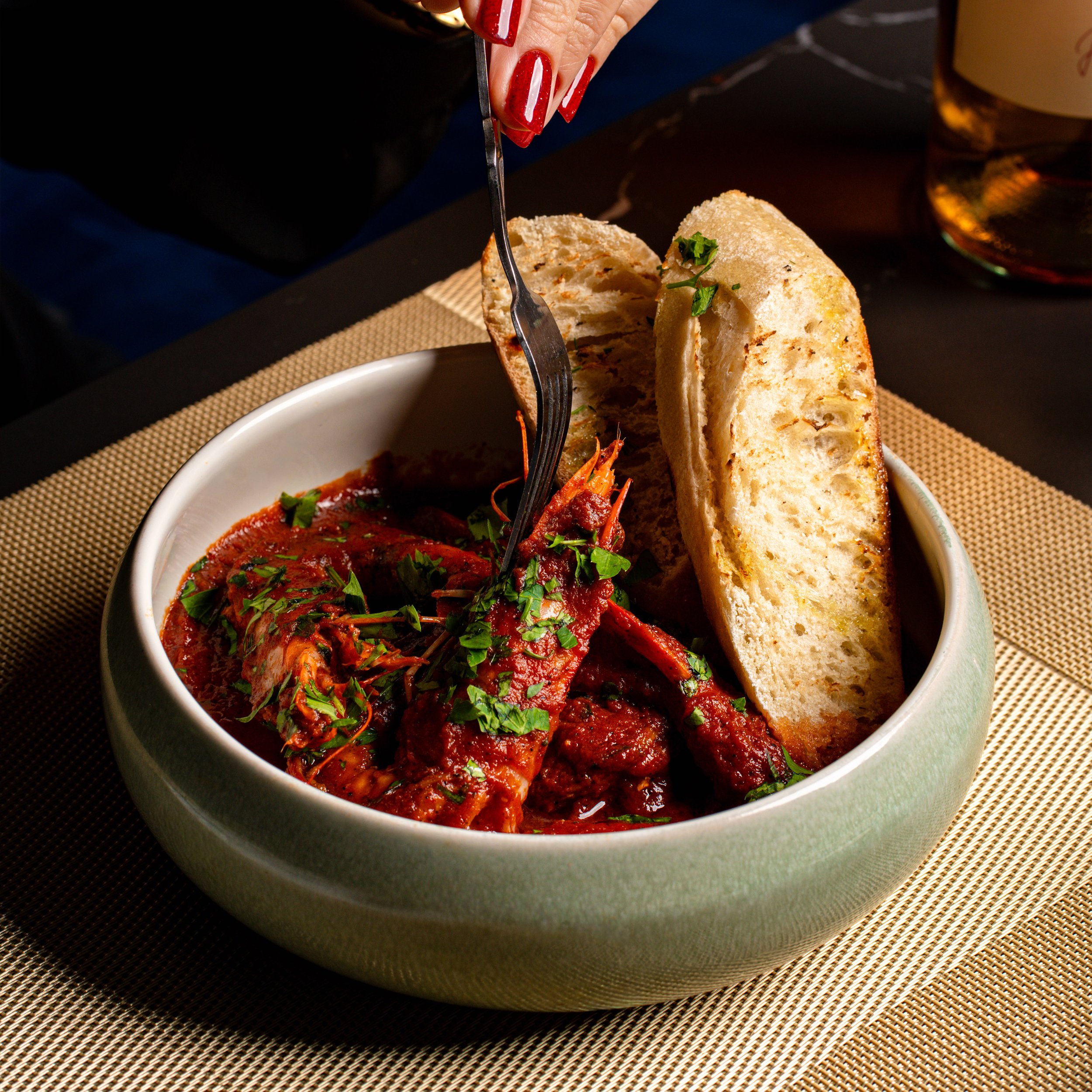 Close-up of a person's hand with red fingernail polish using a fork to cut into bread on a plate of food. The bread is toasted and garnished with parsley, and there is a bowl of tomato-based stew or sauce with herbs underneath. The setting includes a woven placemat and a dark table.