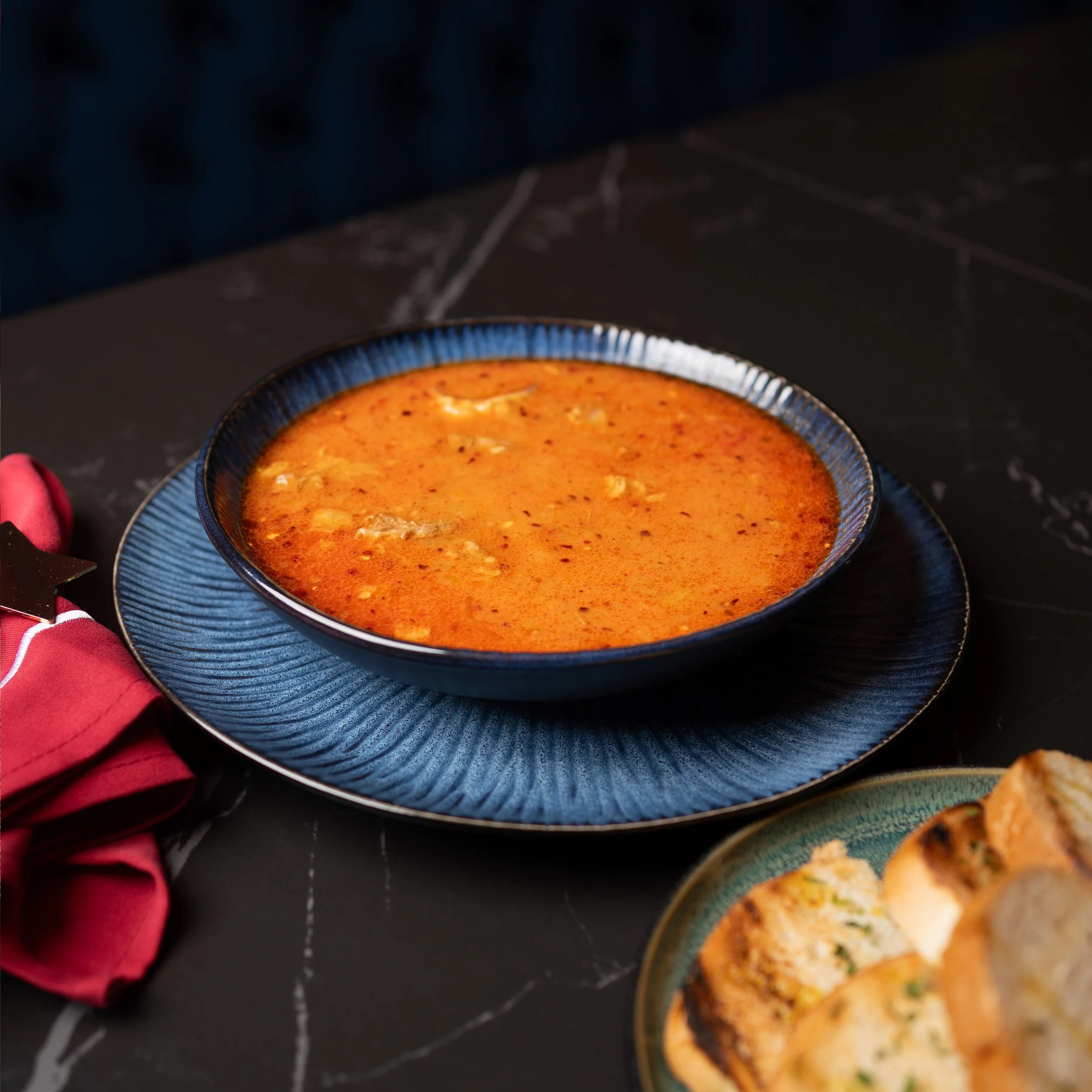 Close-up of a bowl with orange-colored soup or sauce, placed on a dark surface, with a spoon and other bowls partially visible around it.