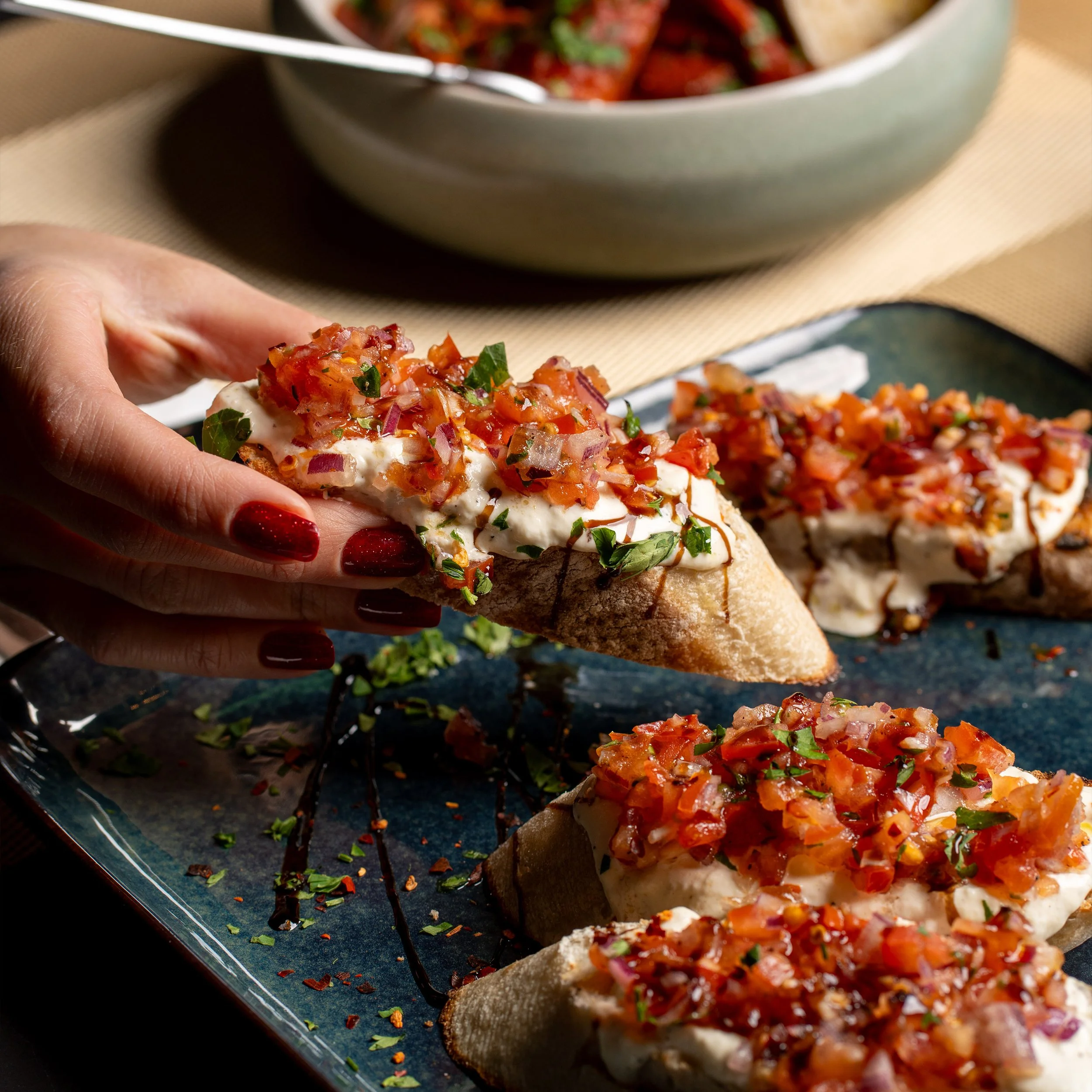 Close-up of a hand holding a slice of pizza topped with chopped tomatoes, onions, and herbs, with a large bowl of similar pizza slices in the background.