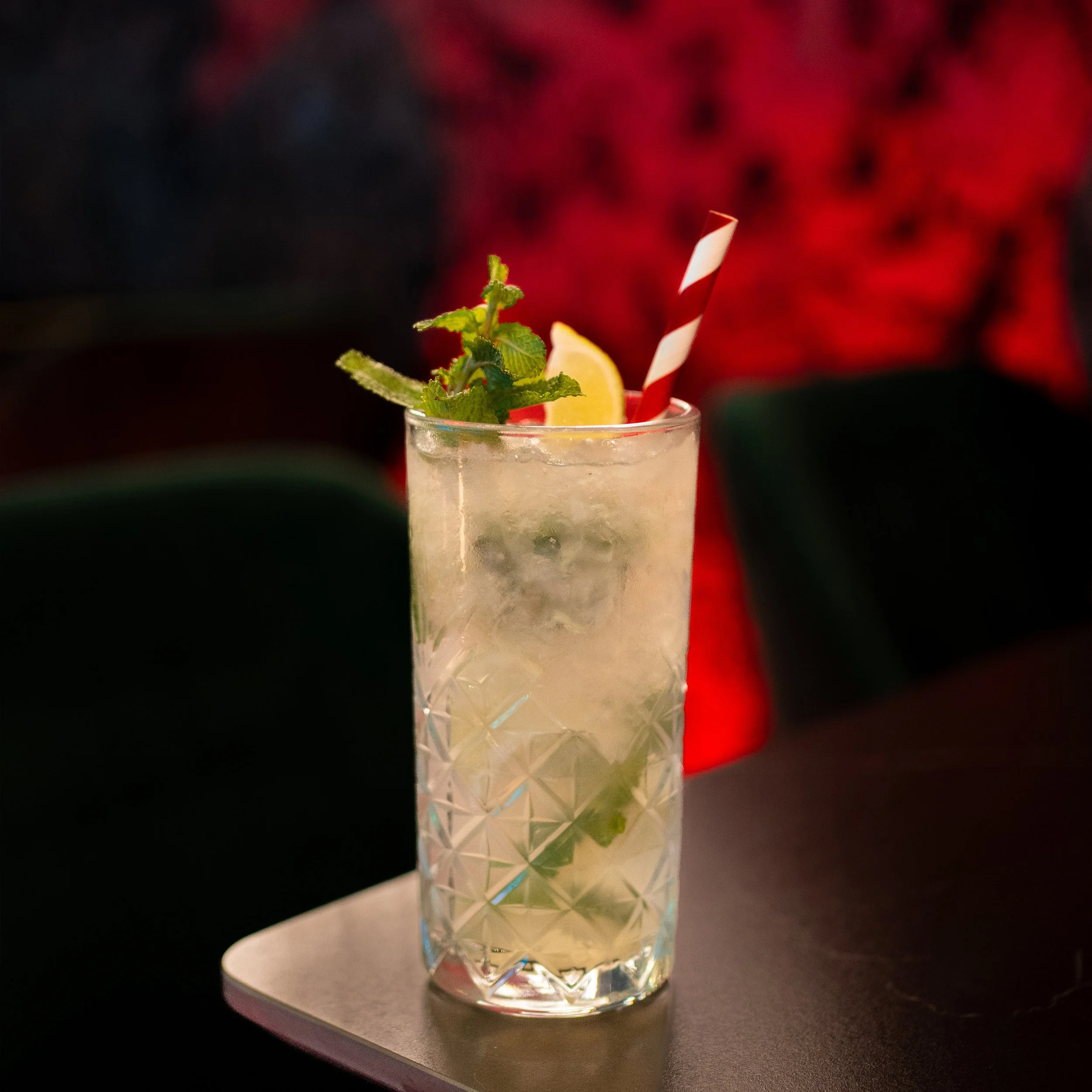 A close-up of a glass containing a beverage with ice, garnished with a slice of lemon, a sprig of mint, and a red and white striped straw, placed on a dark surface with a blurred red background.