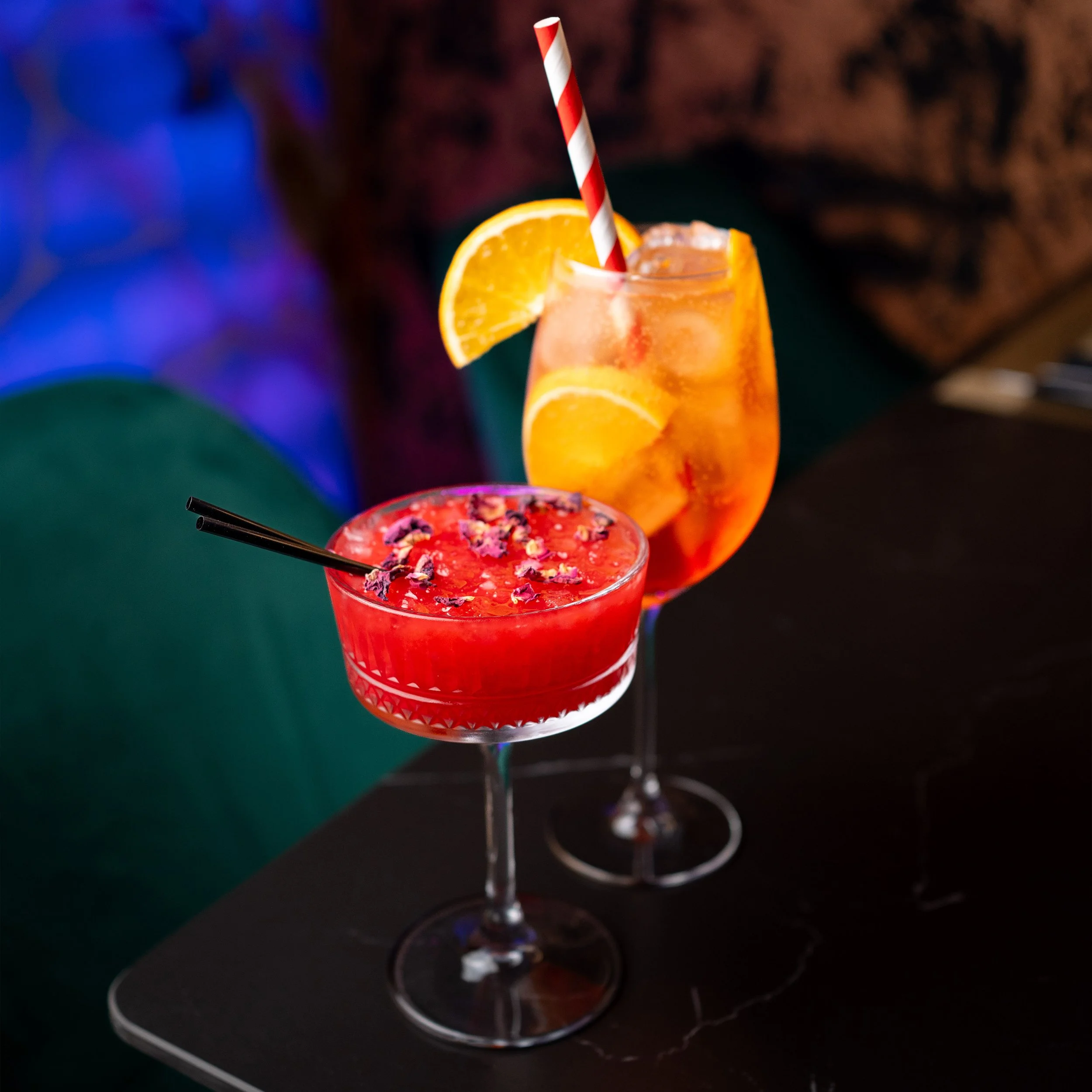 A close-up of two colorful cocktails on a table in a dimly-lit environment. One drink is red with purple flower petals on top, and another is orange with slices of orange and a striped straw. The background is blurred with hints of blue and brown ton