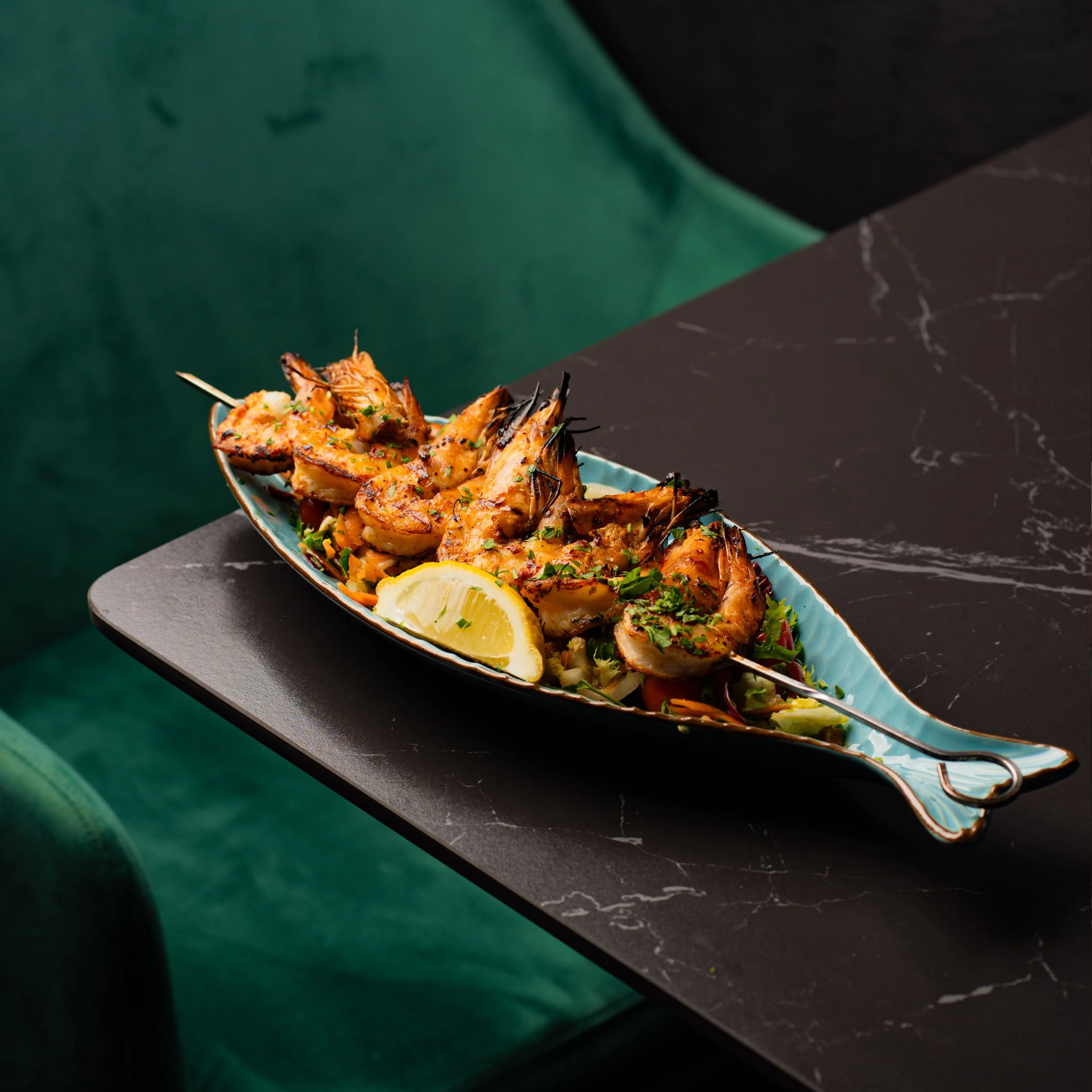 Close-up of a plate of grilled shrimp garnished with herbs, lemon wedges, and vegetables on a dark table with green and black backgrounds.