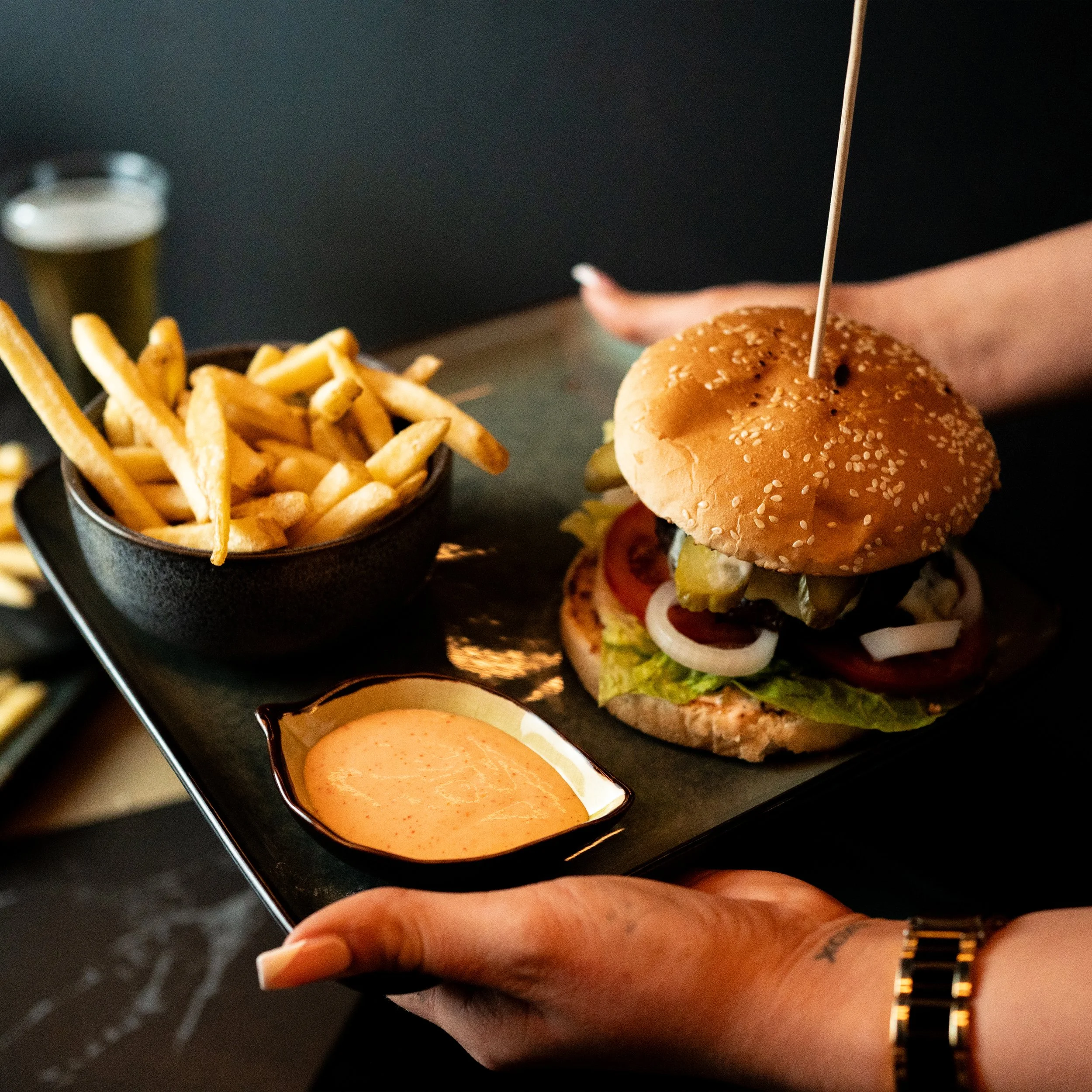 A person's hand holding a smartphone, viewed from above, with a plate of French fries and a burger with a sesame seed bun, vegetables, and sauce, in a restaurant setting.