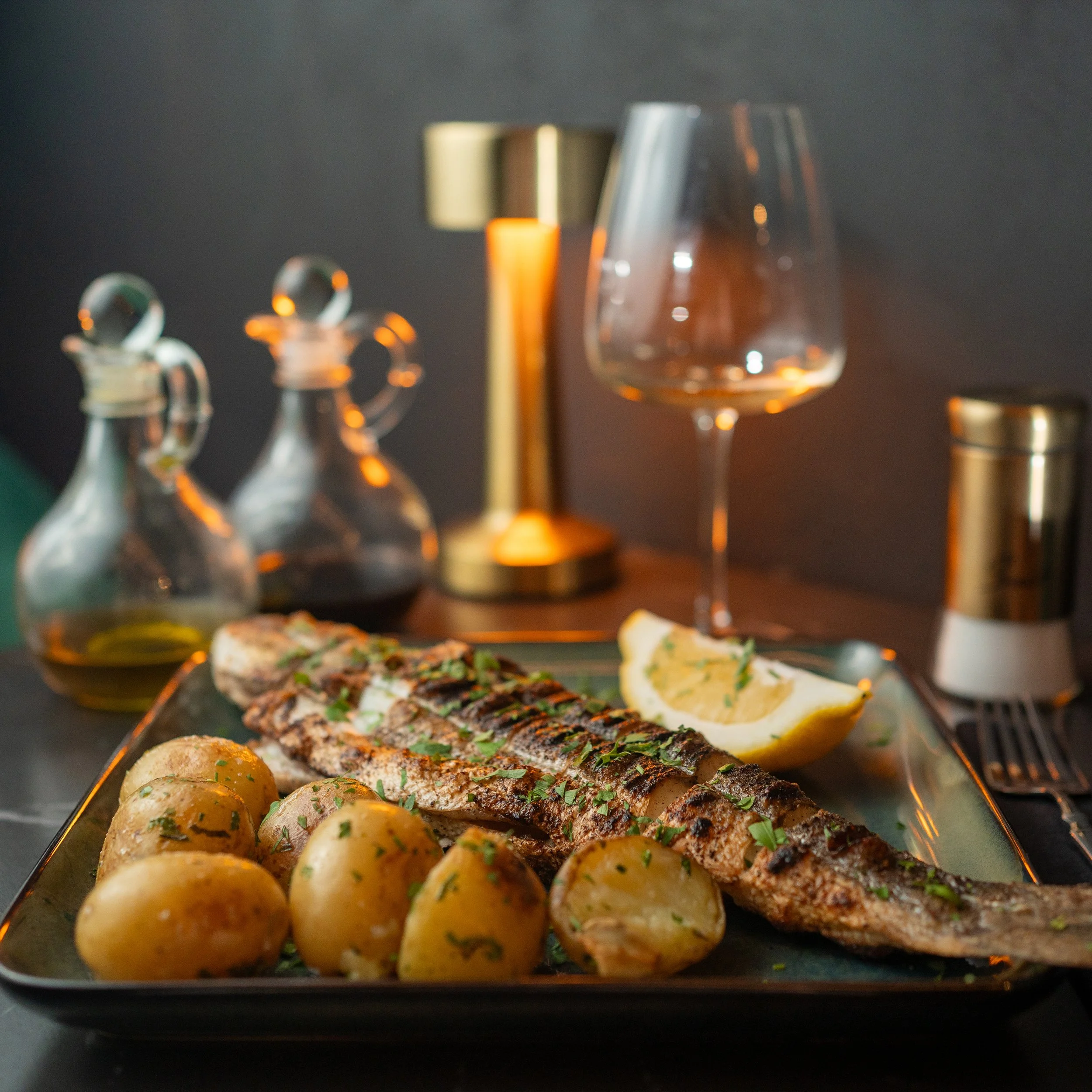 Close-up of a platter with small round roasted potatoes garnished with herbs, sliced fish seasoned with herbs, lemon wedges, and glassware in a dark setting.