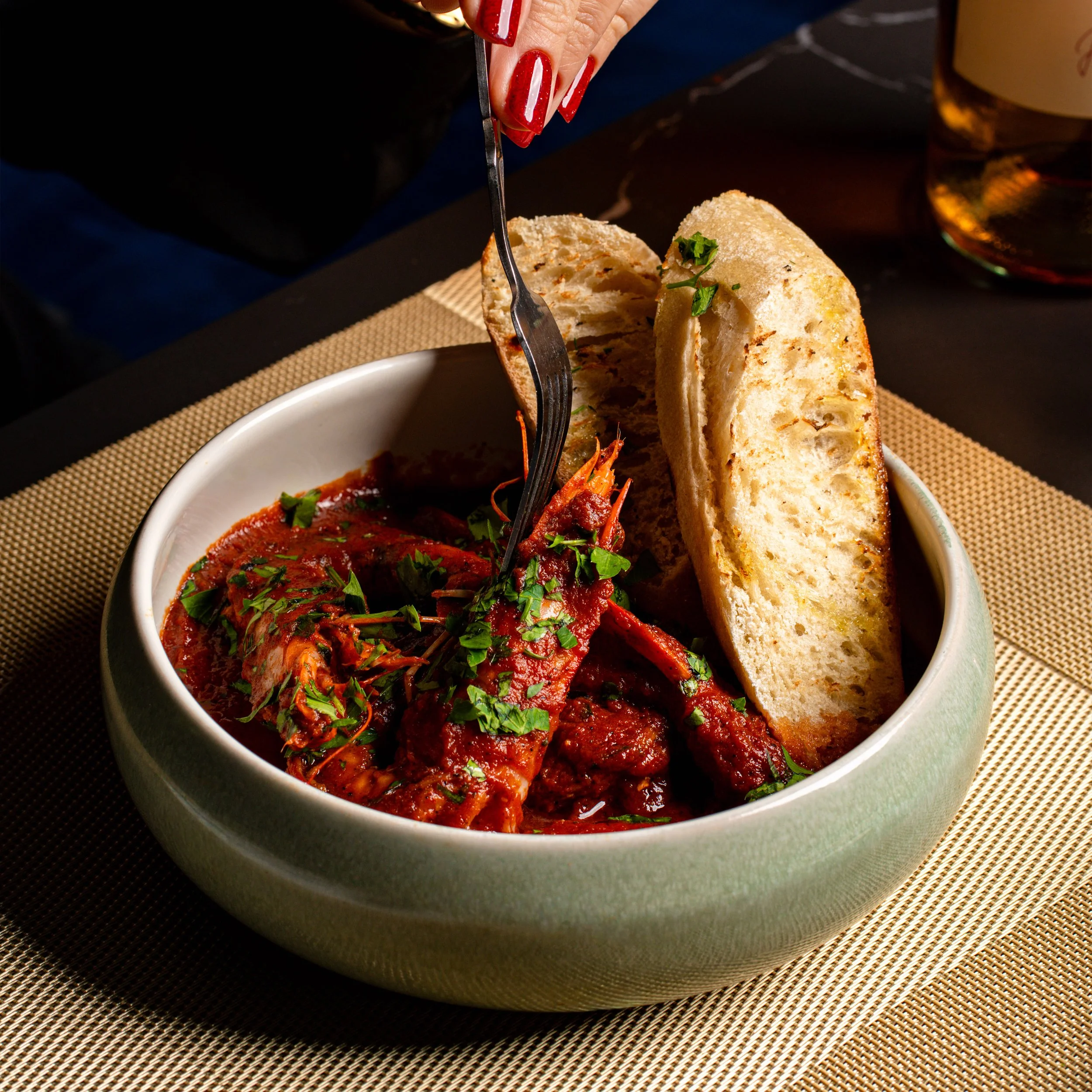 A bowl of spicy seafood stew with tomato sauce and chopped herbs, served with two slices of toasted bread, on a black table with a woven placemat.