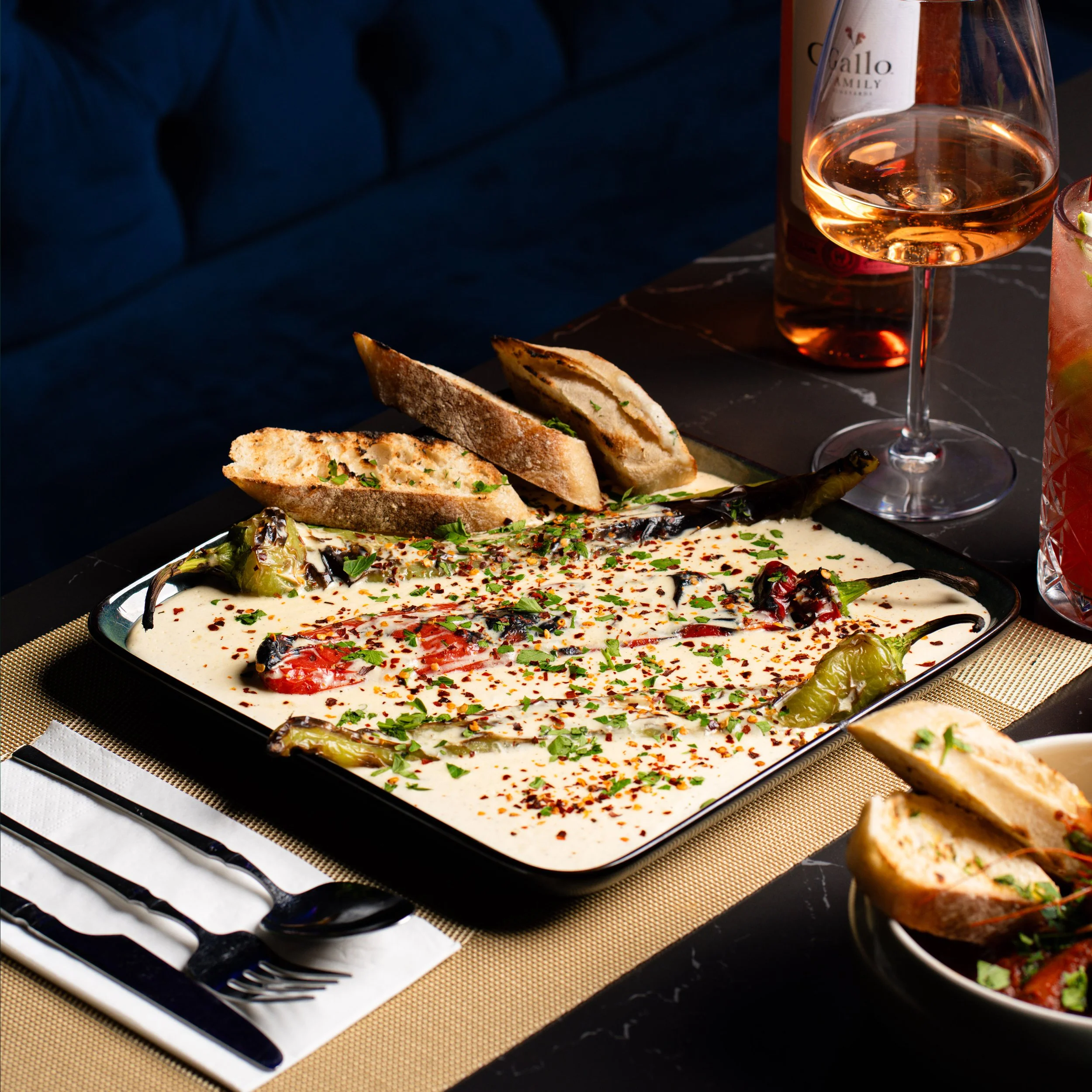A plate of hummus topped with chili flakes and chopped herbs, grilled green peppers, and toasted bread slices with herbs. Behind the plate, there is a glass of rosé wine with bubbles, a glass of pink cocktail with lime slices, and a bottle of rosé wi