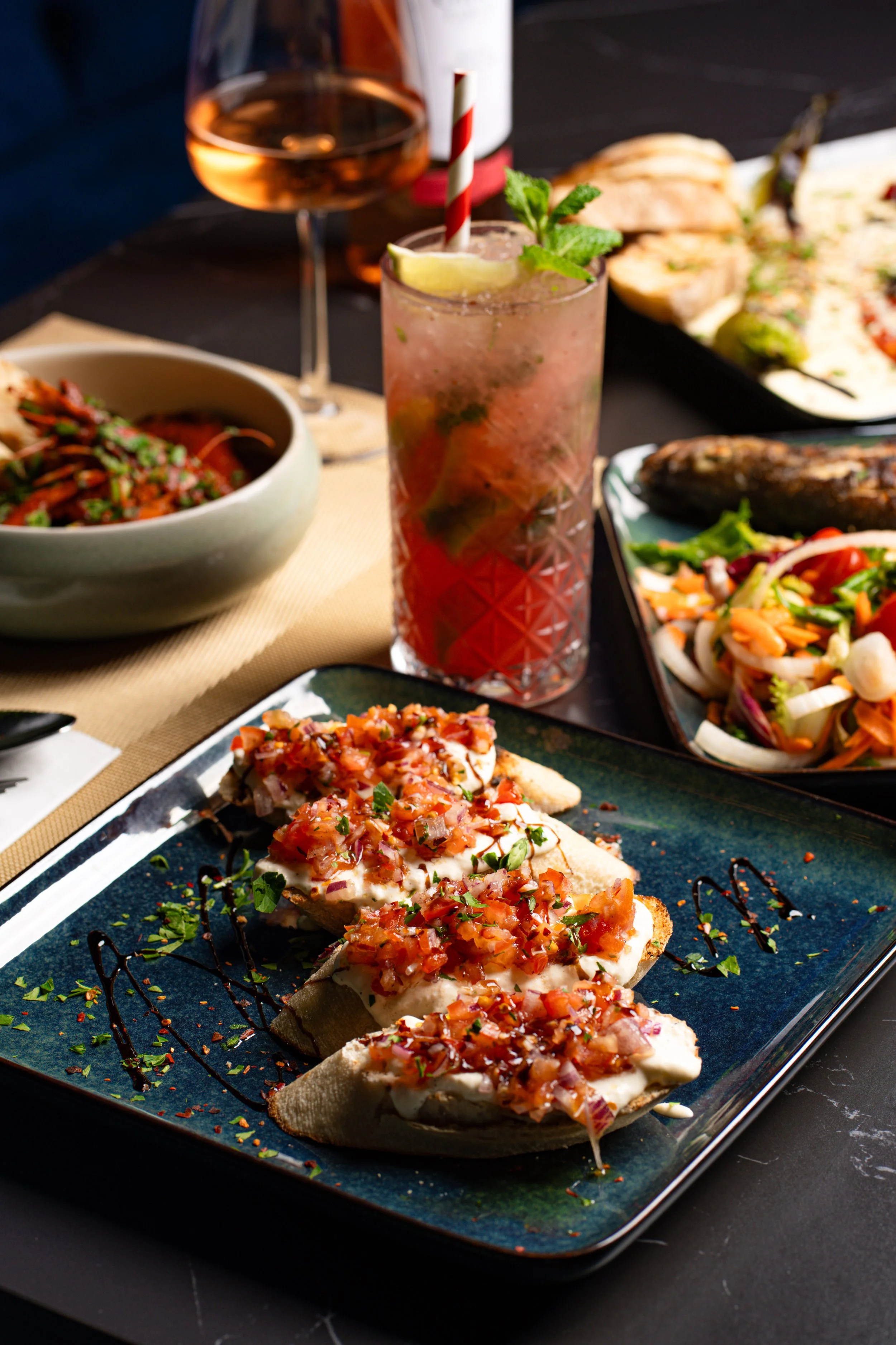 Appetizer of sliced toasted baguette topped with melted cheese and diced tomatoes, garnished with chopped herbs, served on a dark rectangular plate with decorative sauces, with various dishes and drinks in the background.