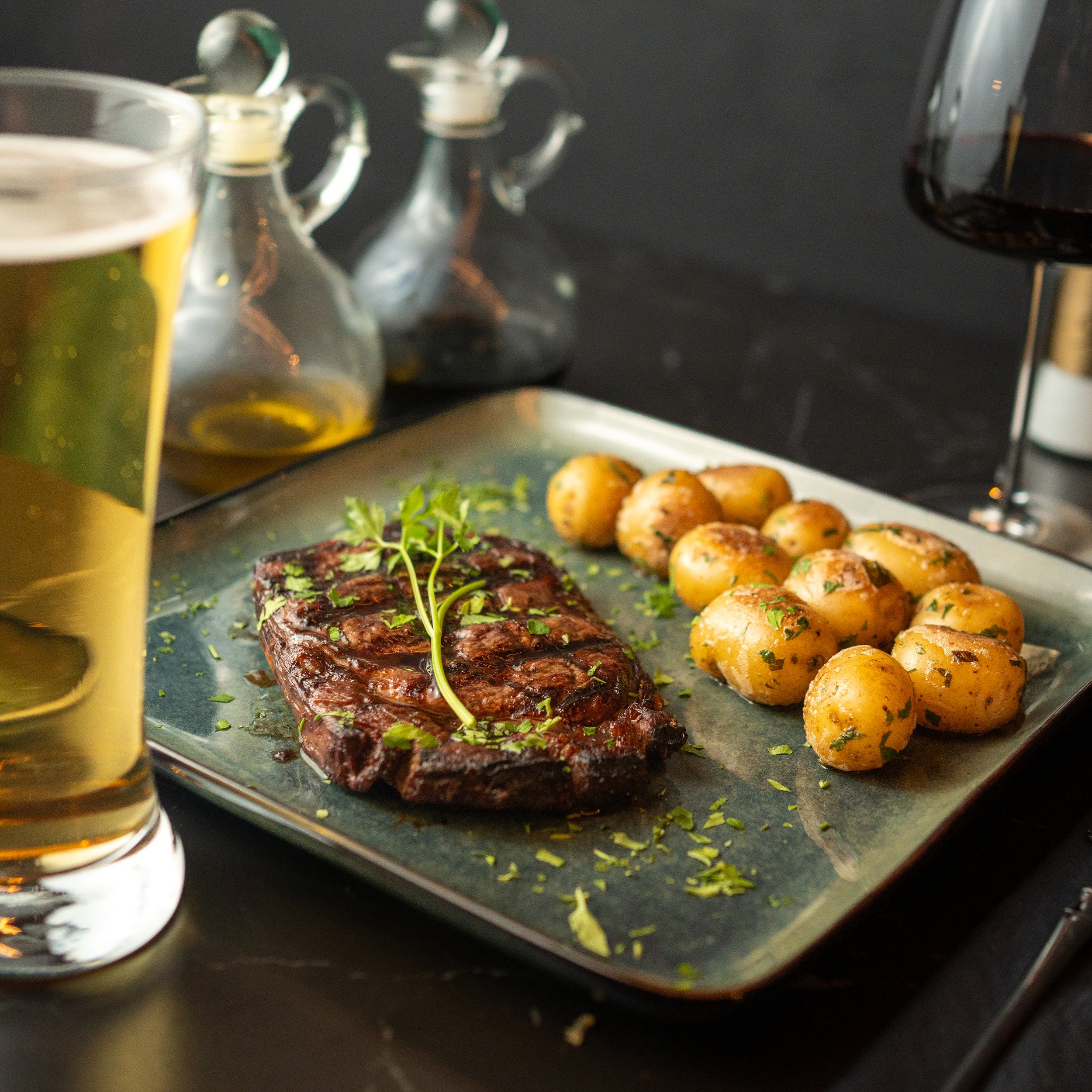 Close-up of a food platter with roasted potatoes and a piece of cooked meat garnished with herbs, with a glass of beer and a dark background.