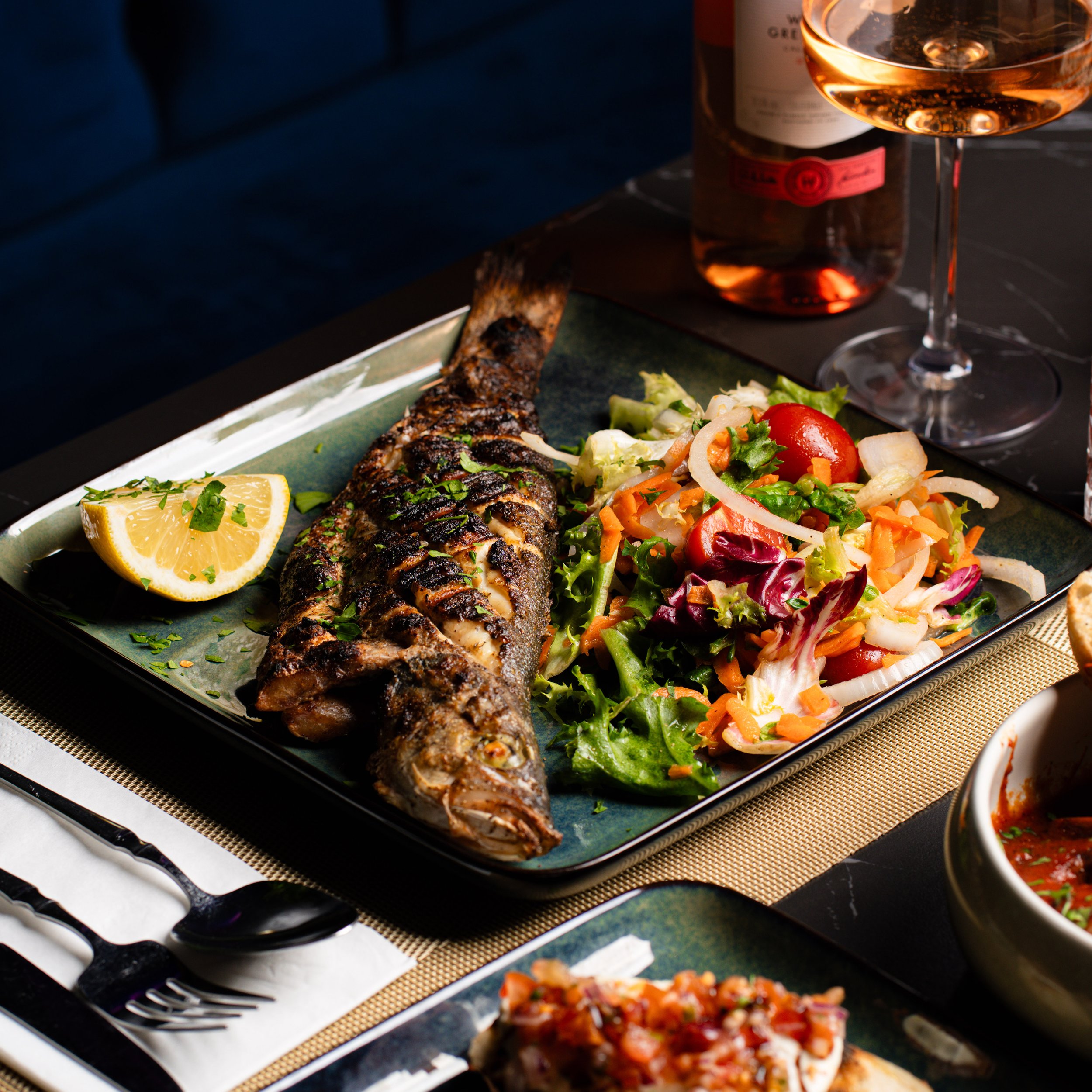 Close-up of a roasted fish fillet garnished with herbs, lemon wedges, and a side salad with cherry tomatoes, lettuce, carrots, and cucumbers on a dark plate, with a glass of rosé wine and a bottle in the background.