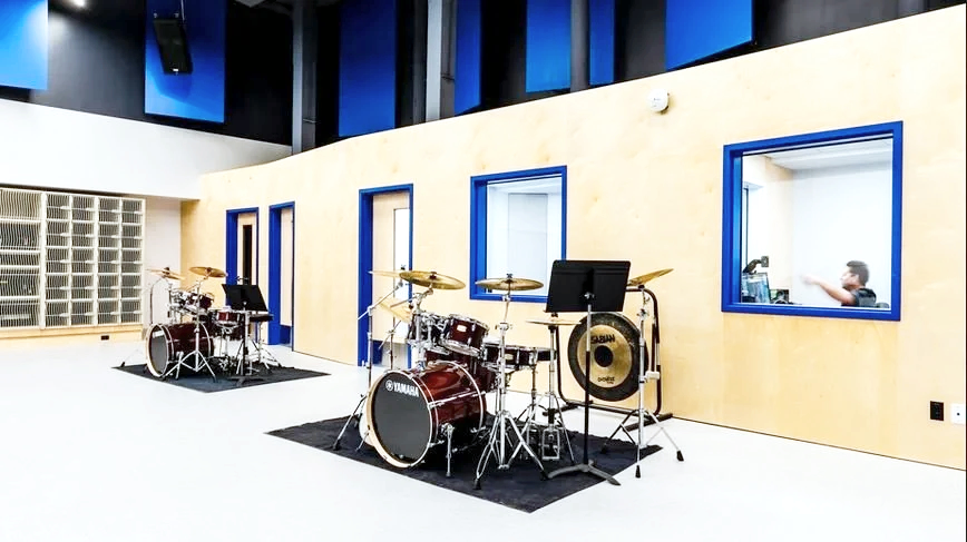 Music recording studio with two drum sets, music stands, and soundproof rooms with blue window frames.