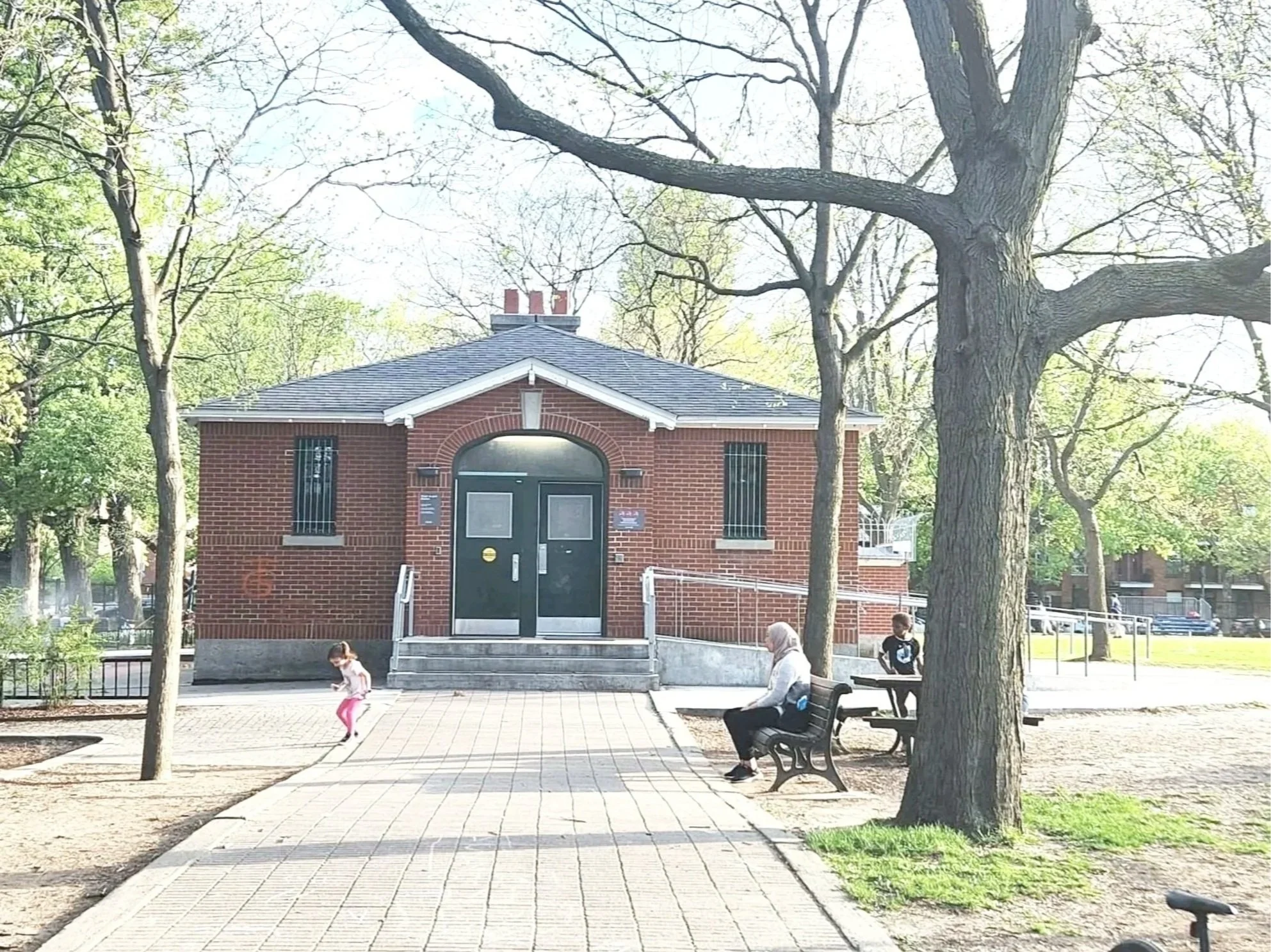 Park scene with children playing, adults sitting on benches, trees, and a small brick building with a black door and barred windows.