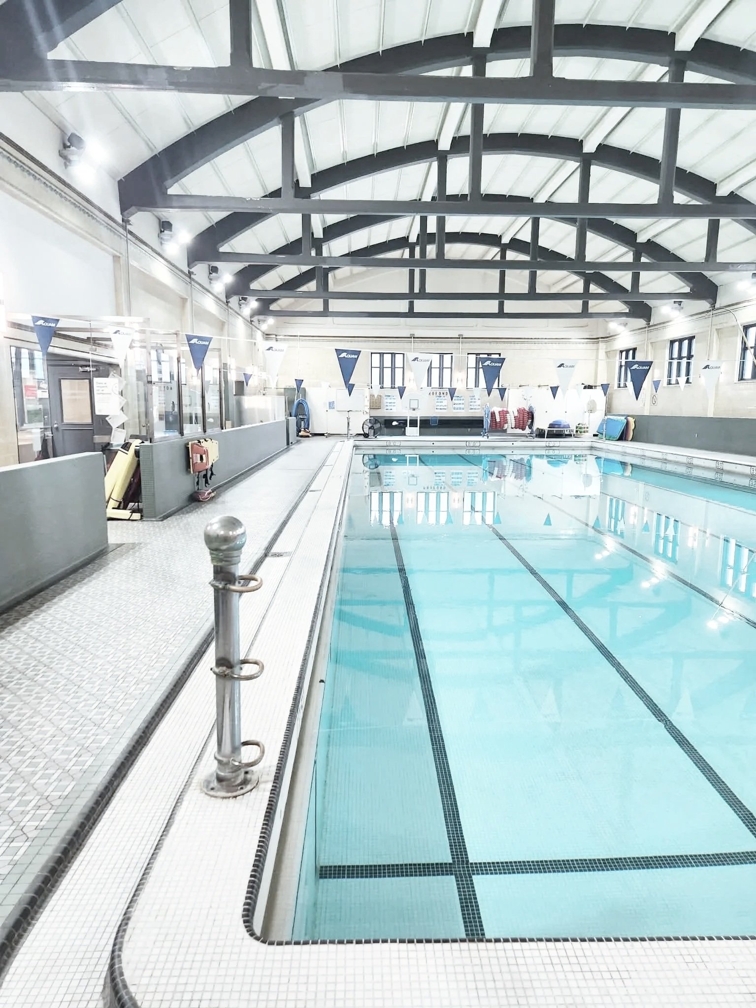 Indoor swimming pool with clear water, surrounded by white tiled floors and walls, and a high arched ceiling with black beams. Pool flags and safety equipment are visible along the side, with windows letting in natural light.