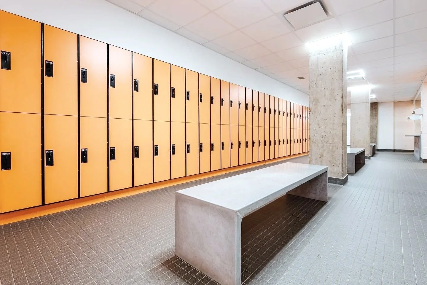 School locker room with yellow lockers, gray tiled benches, concrete pillars, and ceiling lights.