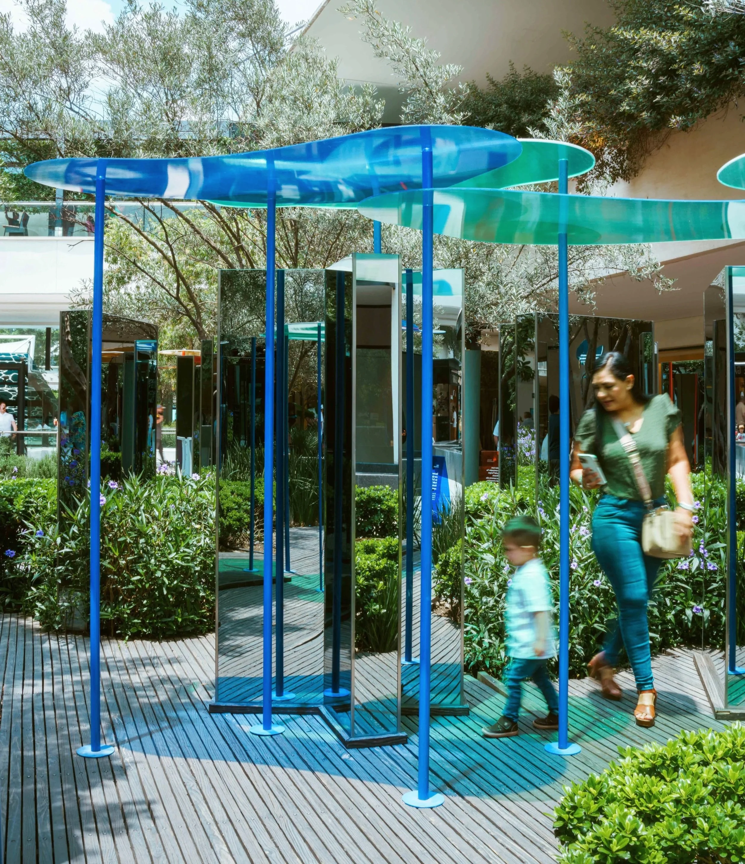 Outdoor scene with two people, a woman and a child, walking on a wooden deck in front of a reflective mirrored installation with tall, slim blue poles and curved, transparent blue and green plastic surfboard-like structures. The background includes trees and modern architecture.