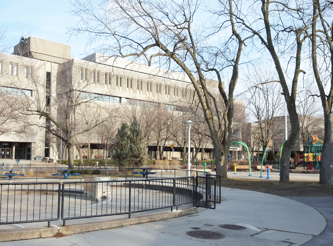 Empty outdoor urban park area with leafless trees, a playground with slides, ride-on equipment, and a large modern beige building in the background.