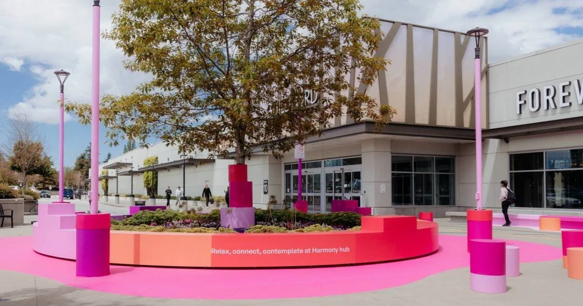 Outdoor public space with a circular pink and orange seating area, pink lamp posts, and trees, outside a shopping mall with people walking by.