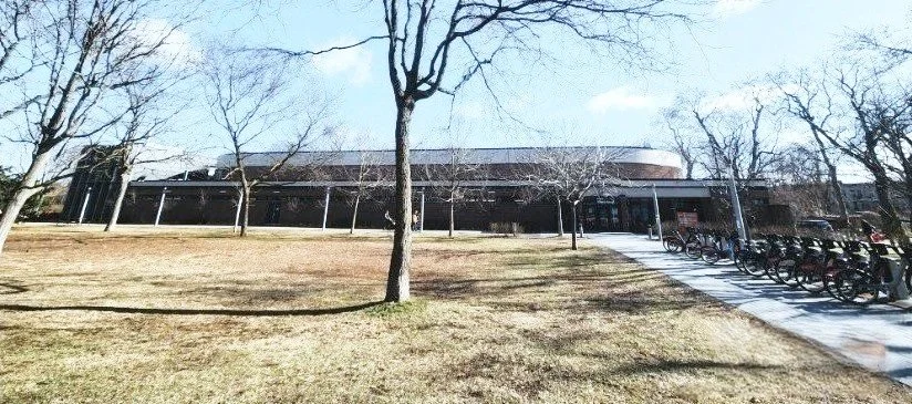 A large, curved building with a sidewalk and bikes parked outside, surrounded by trees with bare branches and a grassy area.