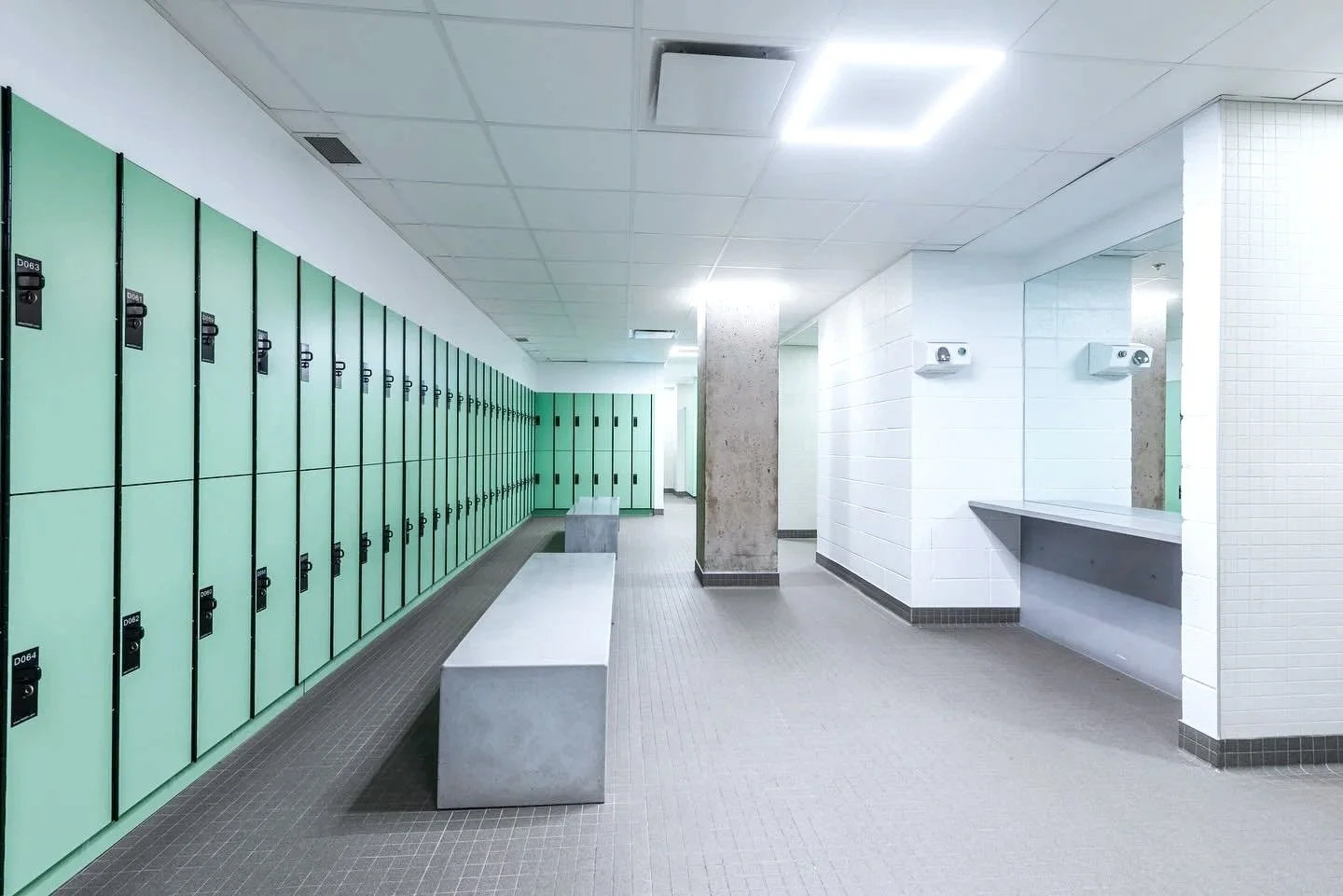 School locker room with green lockers on the left, a concrete bench in the center, and white tiled walls with shelves on the right.