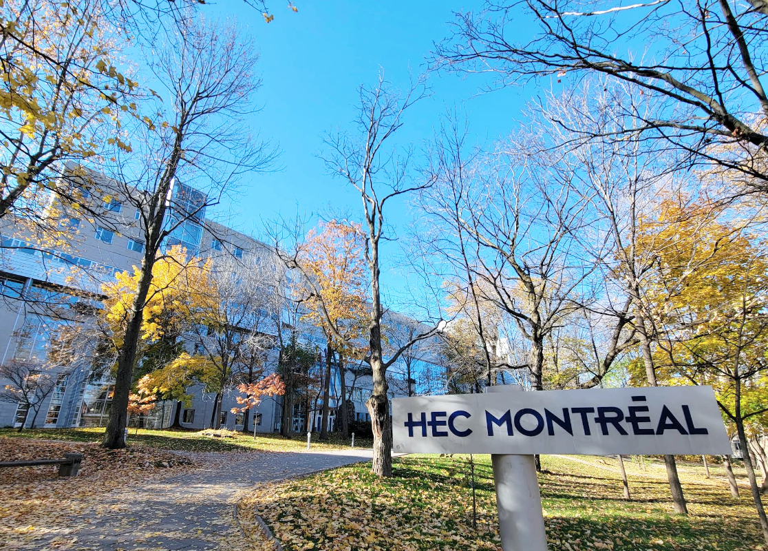 Beautiful autumn scene at HEC Montreal campus with trees shedding yellow leaves, a clear blue sky, and a sign reading 'HEC Montréal'.